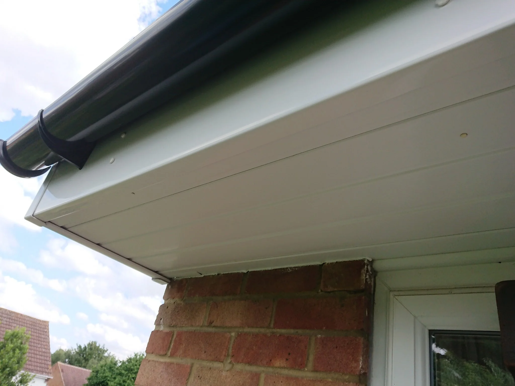 Close-up of the underside of a house's roof eaves showing white siding, brick wall, and part of a window with a white frame.