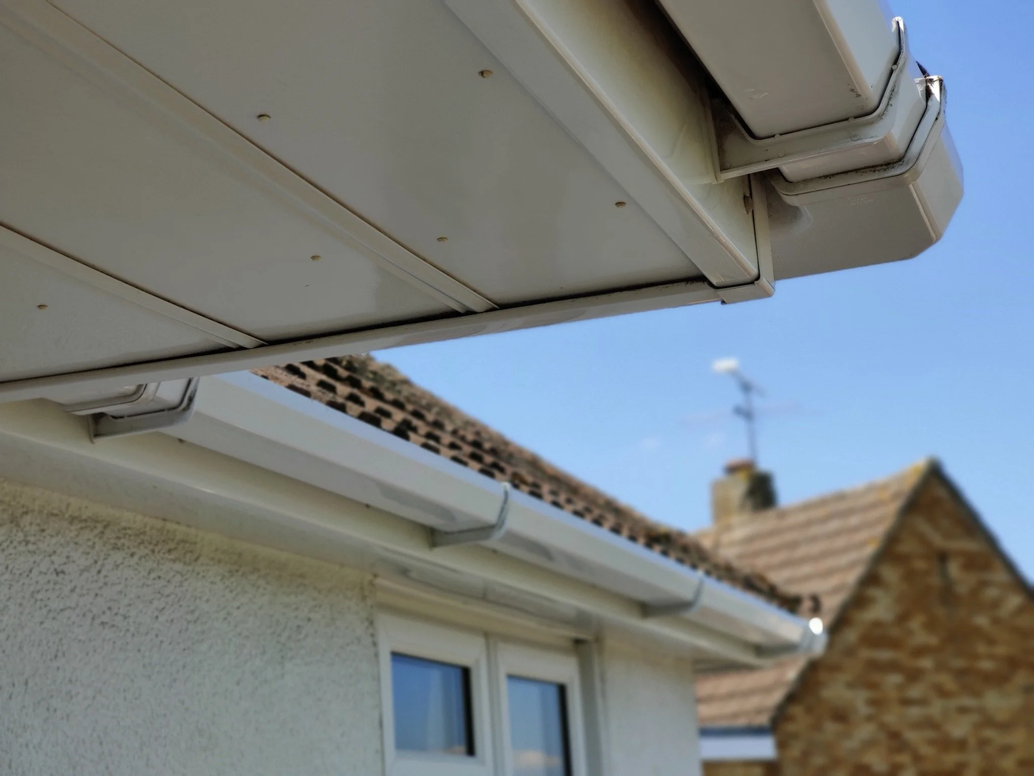 Close-up of house gutters and roof eaves against a blue sky, with part of a neighboring house and chimney in the background.