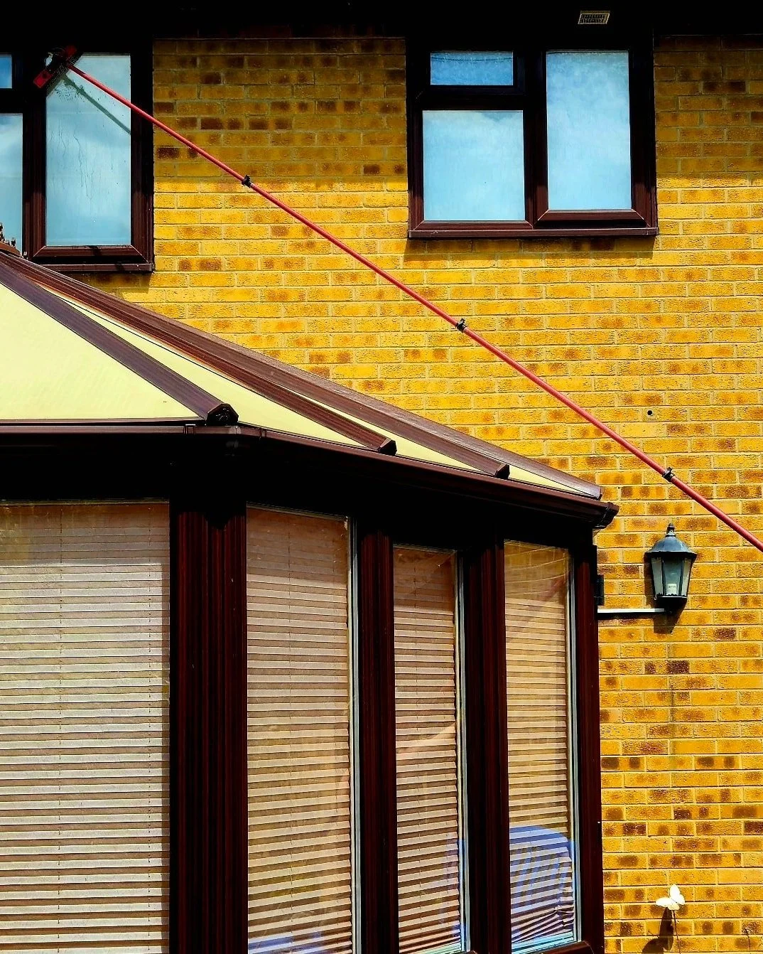 Close-up of a yellow brick house wall with three windows, an exterior light fixture, and a temporary |multi-purpose| red pole extending diagonally across the window area.
