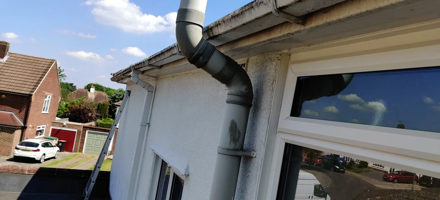 View of the side of a house with a white textured wall, a large plastic gutter pipe, and two windows with white frames, one with a small section of an awning. The reflection of the sky and clouds is visible on the window glass. In the background, res