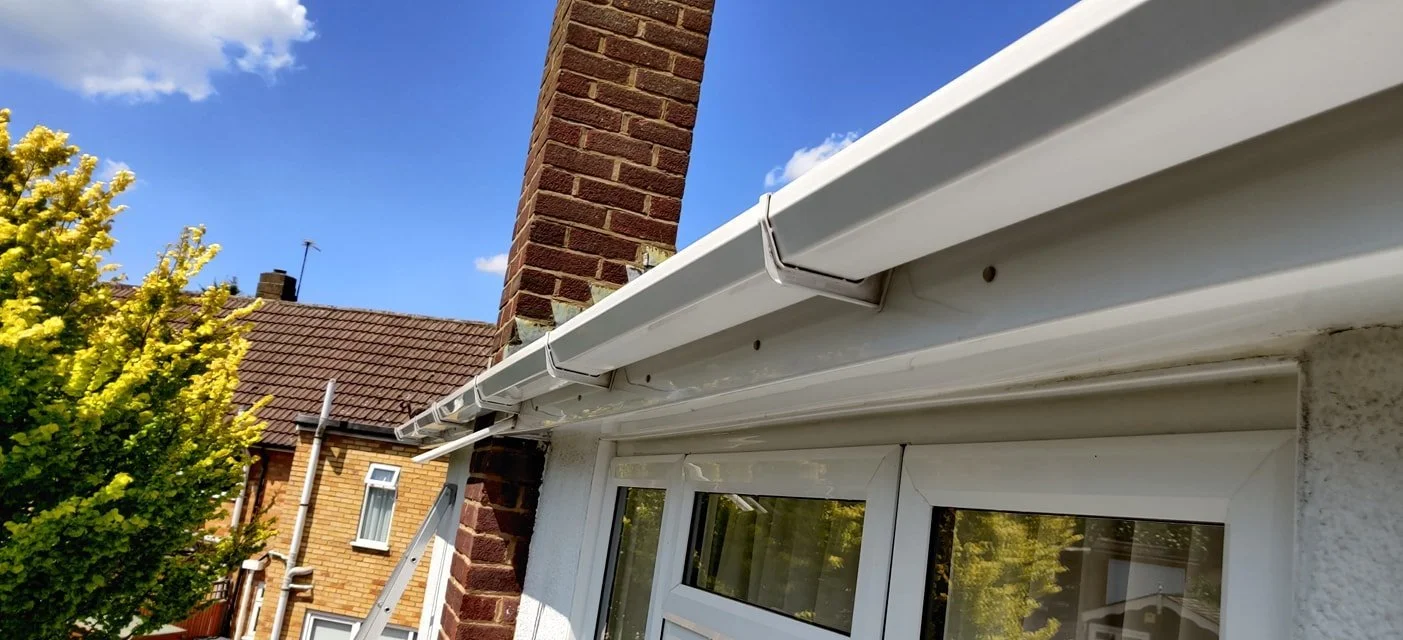 Close-up of a white window with a gutter system on a brick house, with a background of neighboring brick houses and a blue sky with some clouds.
