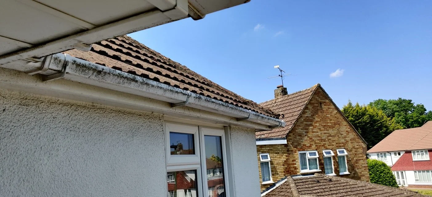 Close-up view of the corner of a house's roof and exterior wall with a small window, neighboring houses, and a clear blue sky in the background.