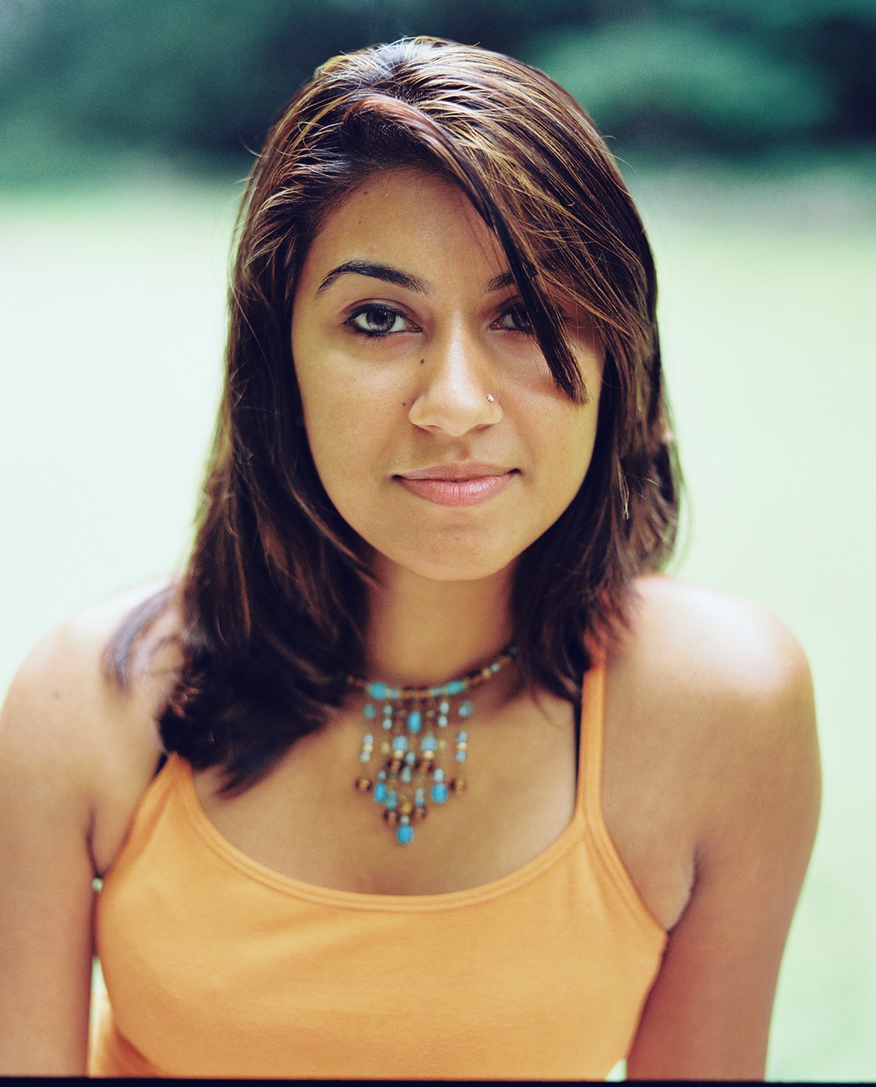 Portrait of a young woman with dark wavy hair wearing an orange tank top and turquoise jewelry, smiling subtly, outdoors with a blurred green background.