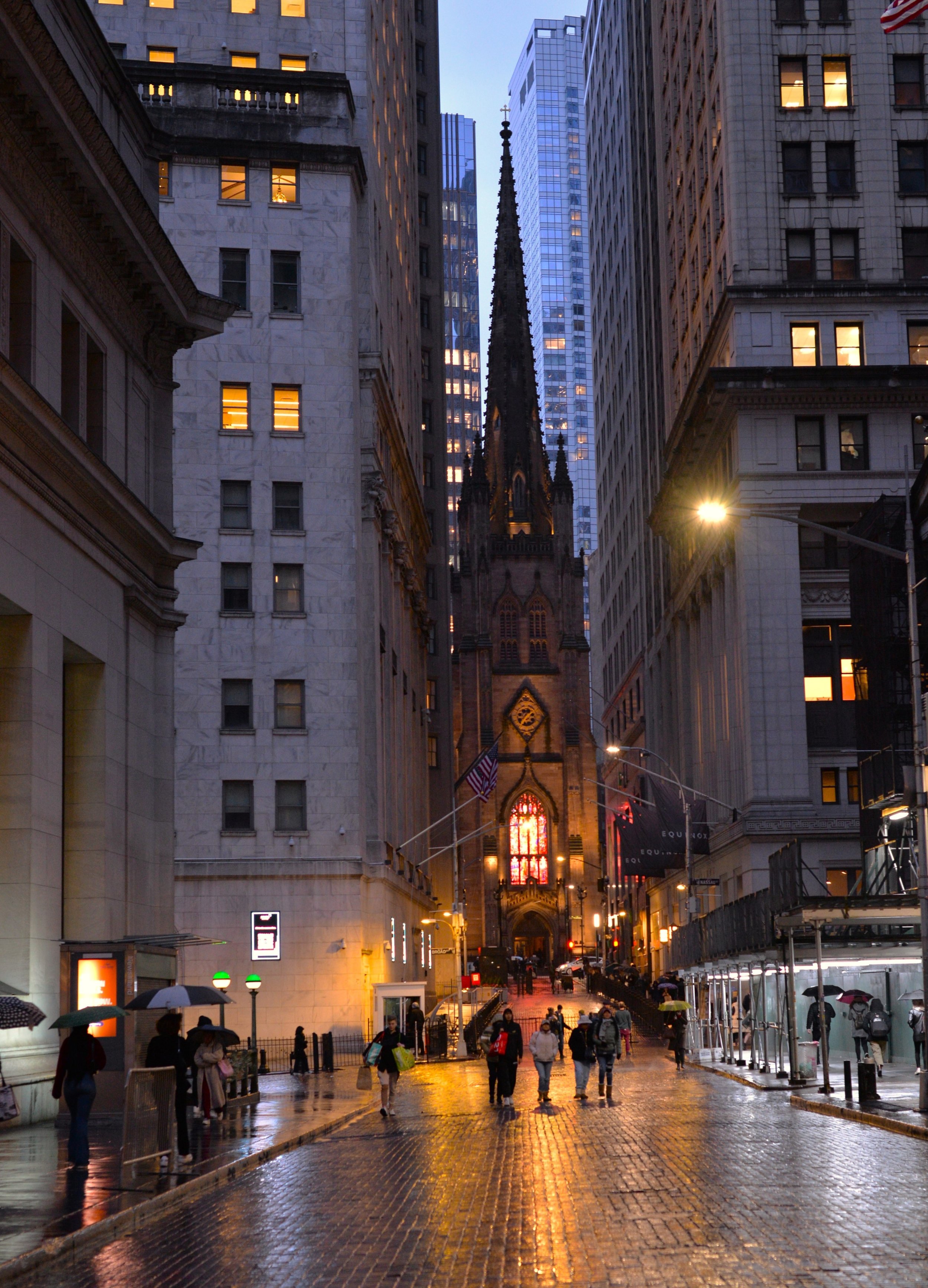 City street scene during evening with wet cobblestone road reflecting streetlights, pedestrians with umbrellas, tall buildings, and a church with stained glass window in background.