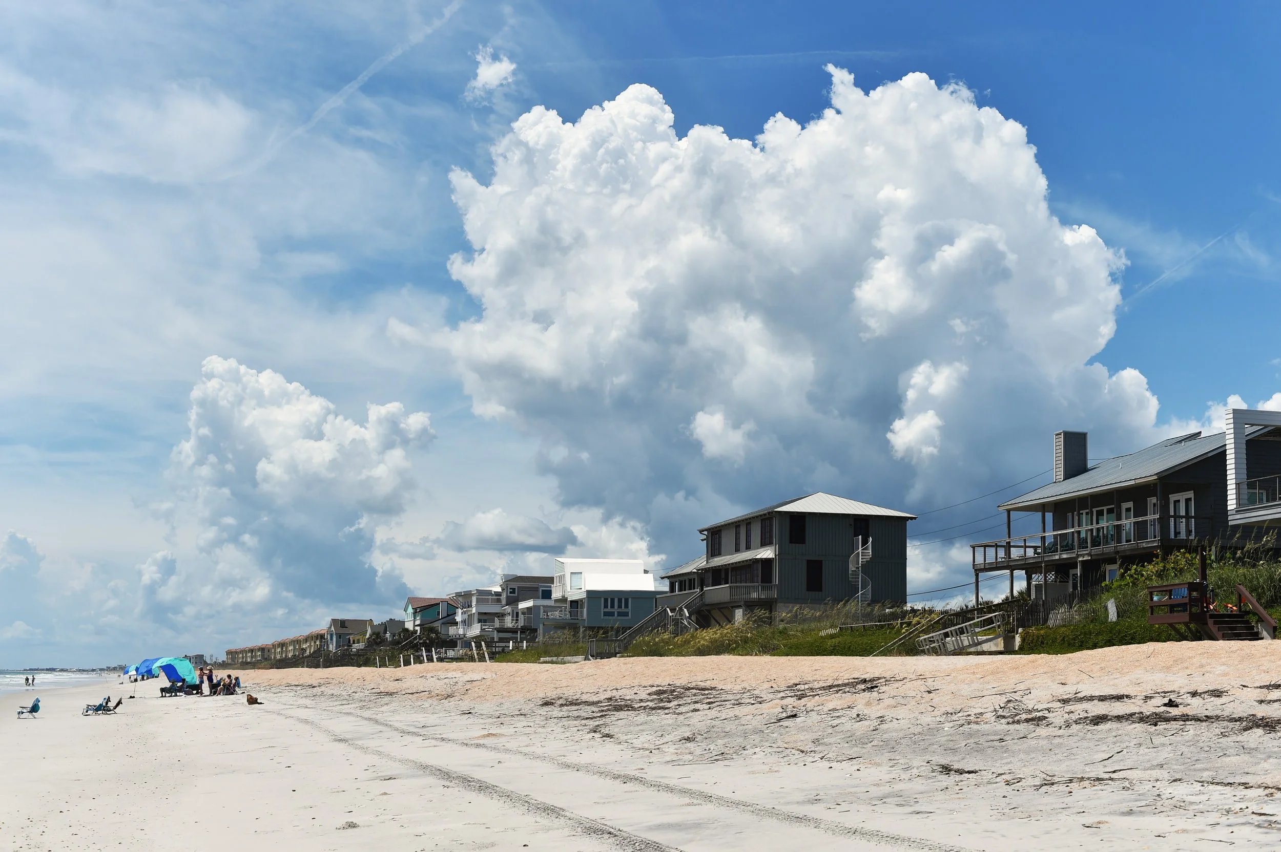 Beach scene with modern houses along the shoreline, cloudy sky, and people relaxing under umbrellas.