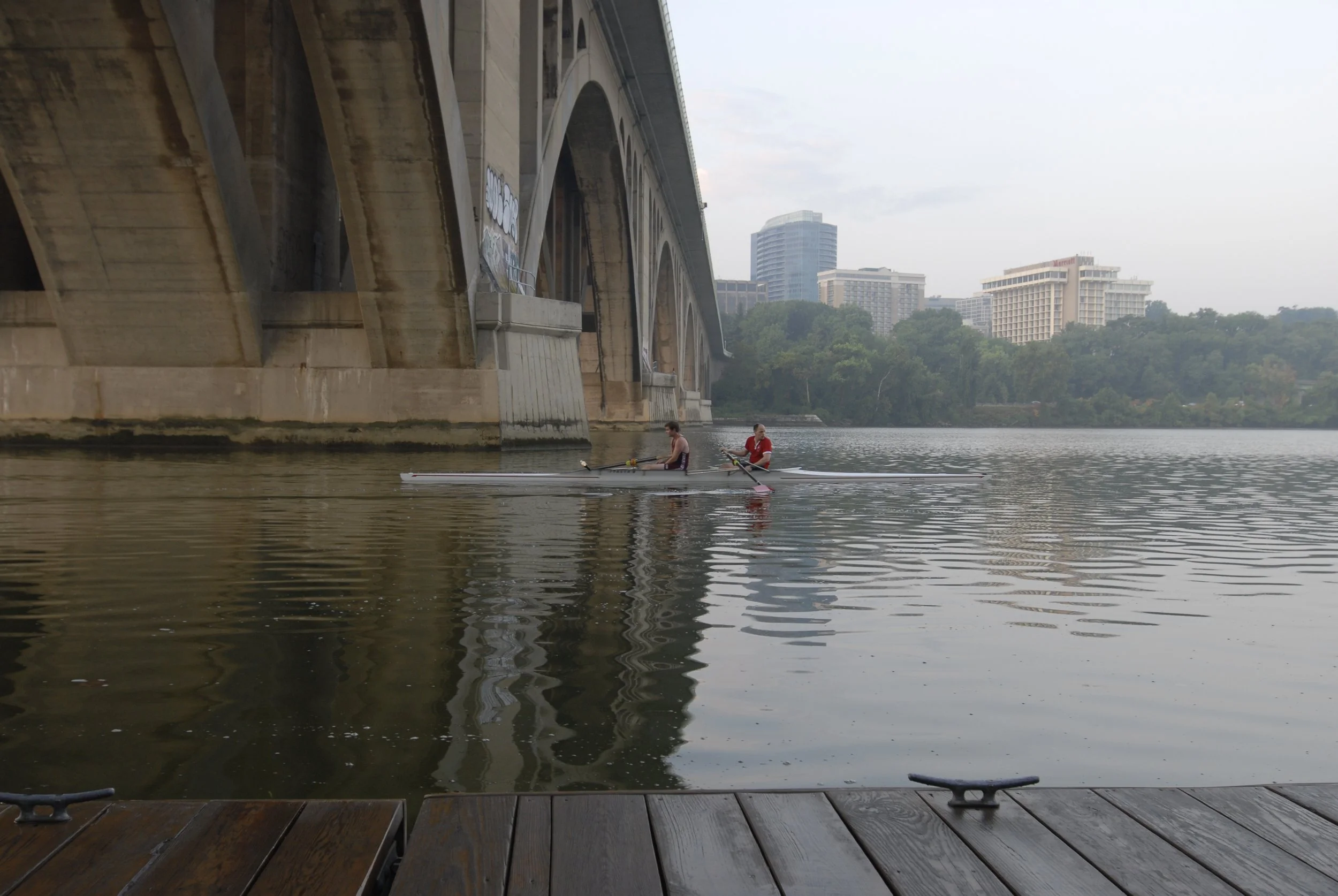 Two people paddling a canoe or kayak on a calm river with buildings and trees in the background, under a bridge.