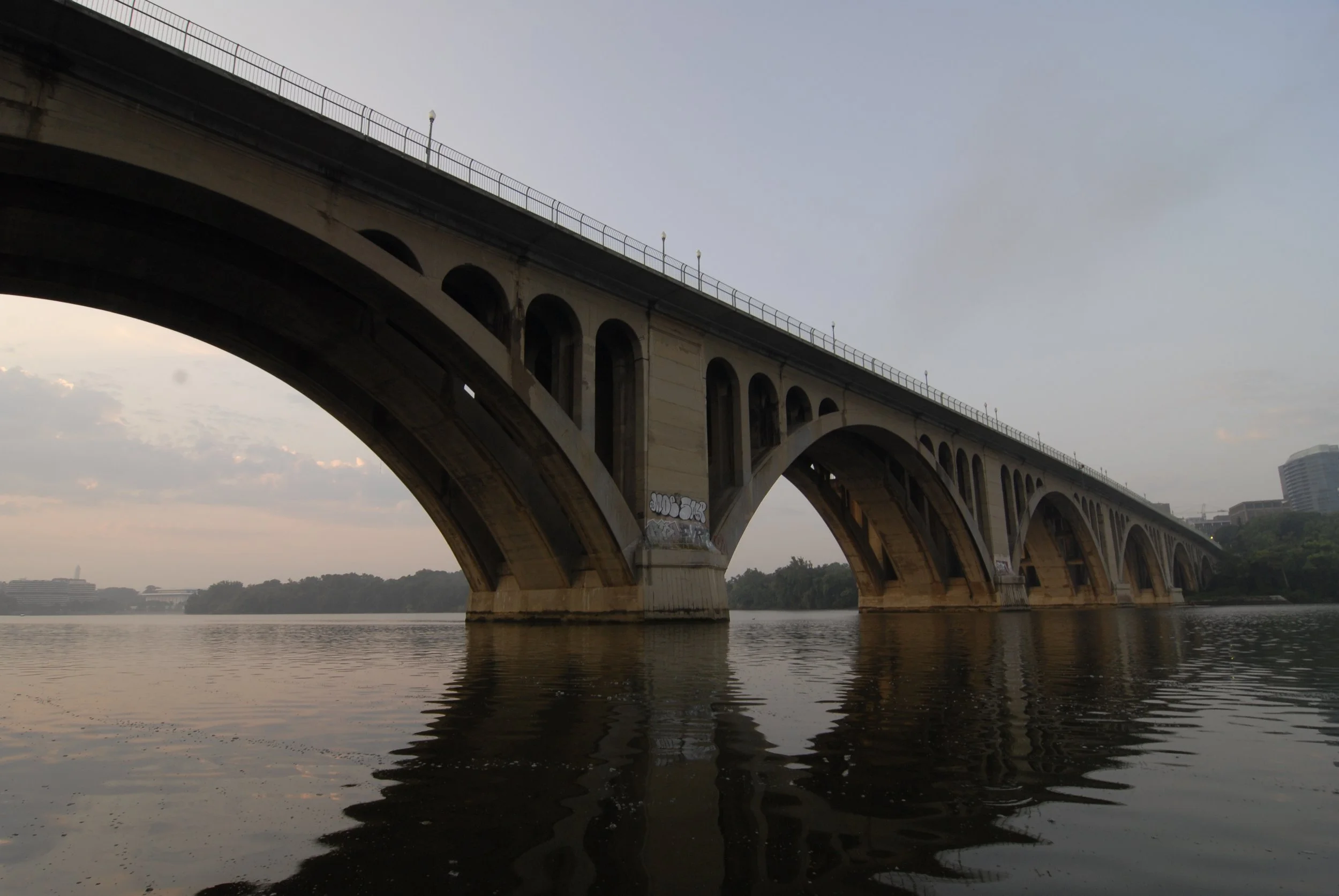 A large concrete bridge with arches over a body of water, with a city skyline and trees in the background, under a cloudy sky.