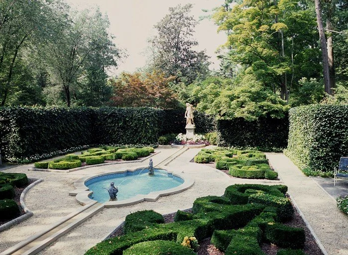 A landscaped garden with a fountain, a statue, well-trimmed bushes, and trees.