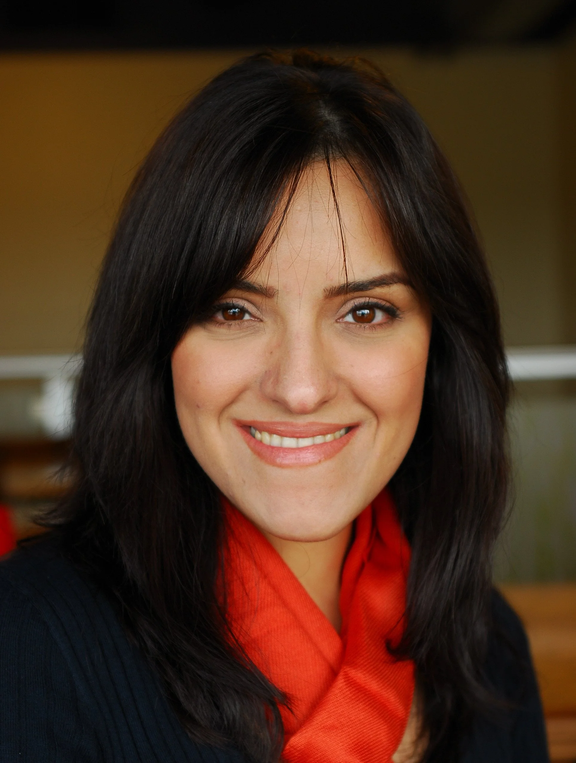 A woman with dark brown hair, brown eyes, and a warm smile, wearing a red scarf and a black sweater, in an indoor setting.