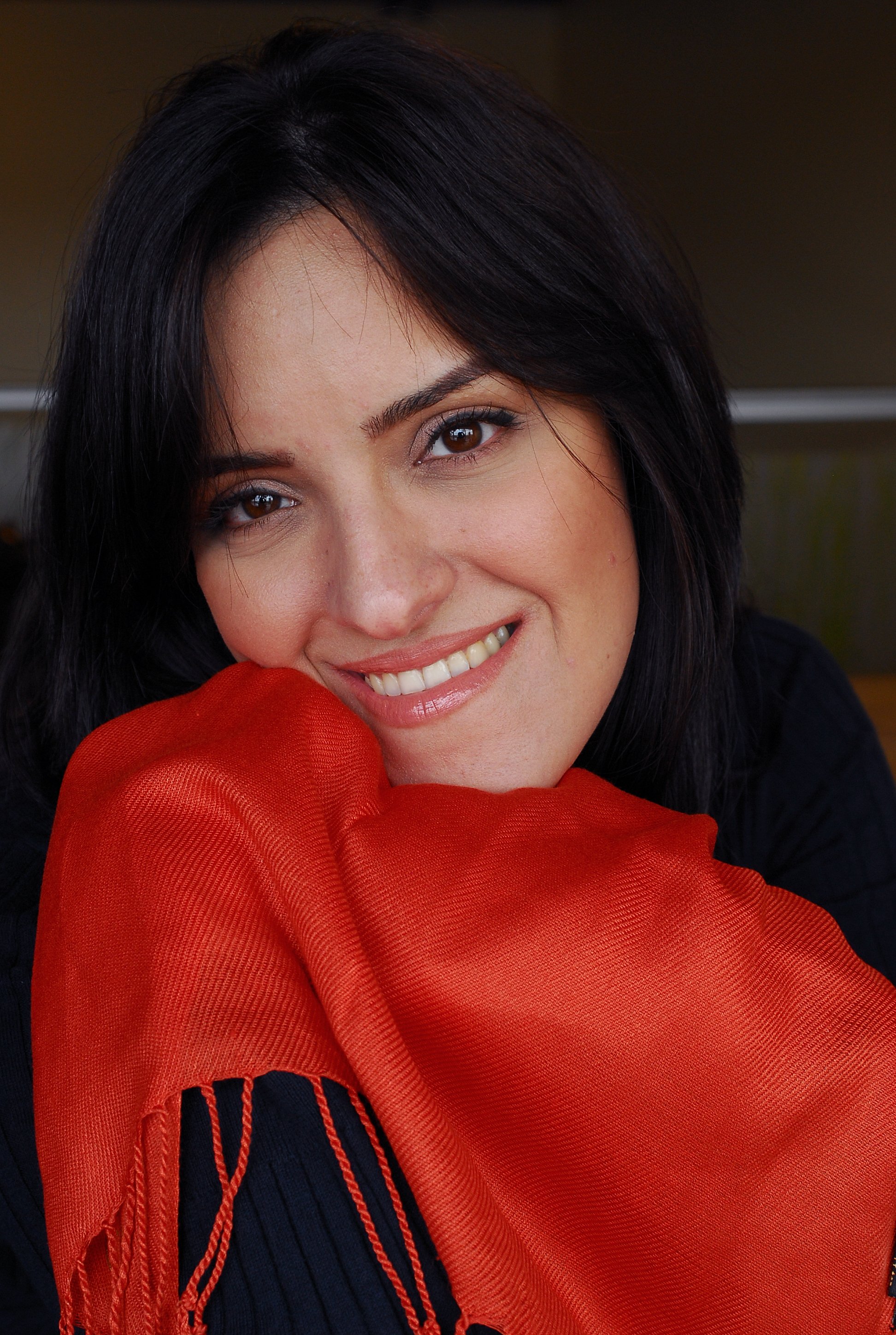A woman with dark hair and brown eyes smiling, resting her face on a red scarf she is holding, with a dark background.
