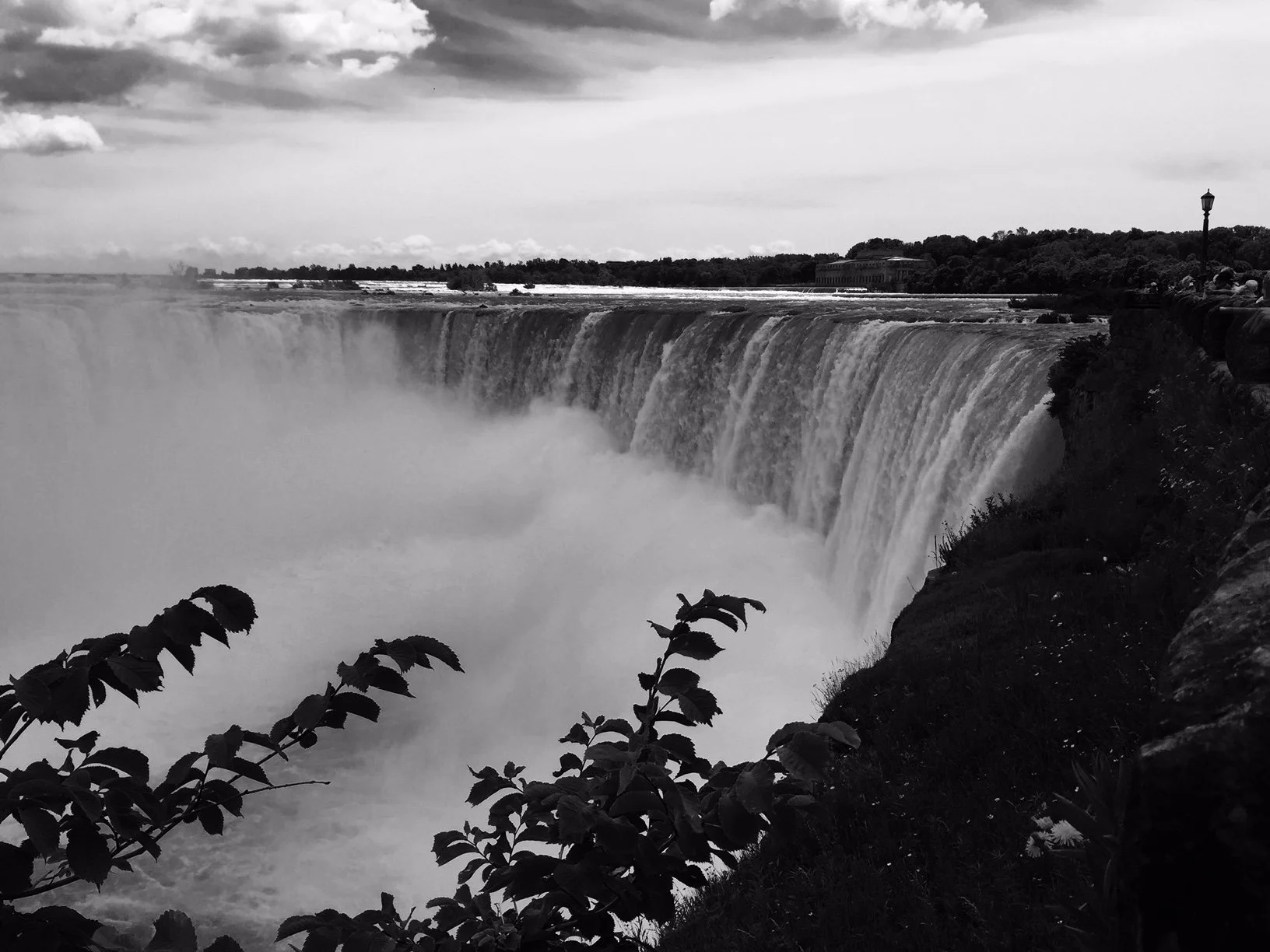 A black and white photo of Niagara Falls, showing cascading water with mist rising from the falls. There are plants in the foreground and a viewing area with a railing and some street lamps on the right side.