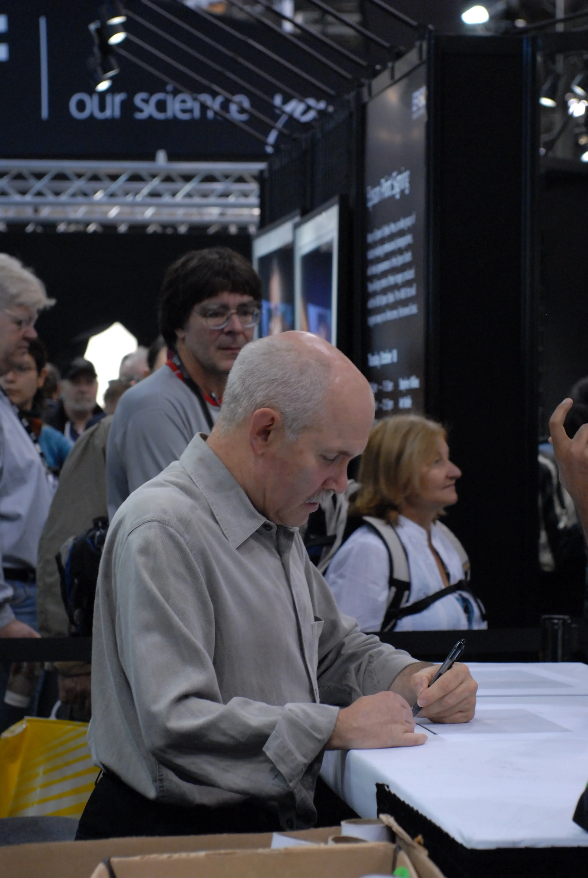 A man with gray hair and a beard wearing a gray shirt is sitting at a table, writing with a pen. Behind him is a crowd of people, some looking at the black display boards. The setting appears to be an event or conference, with a sign in the backgroun