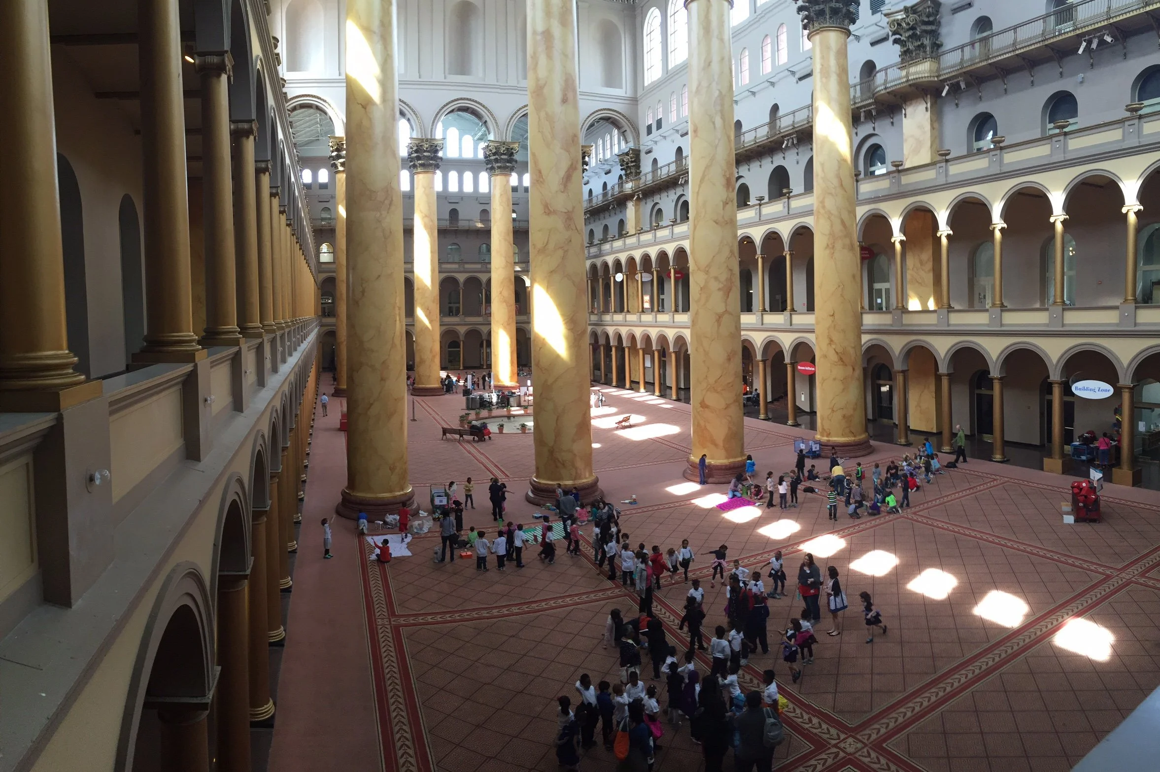 Interior of a grand building with tall, marbled columns and multiple levels with balconies. There are many people, including children, gathered on the ground floor, some sitting on mats. Bright sunlight streams through large arched windows, illuminat