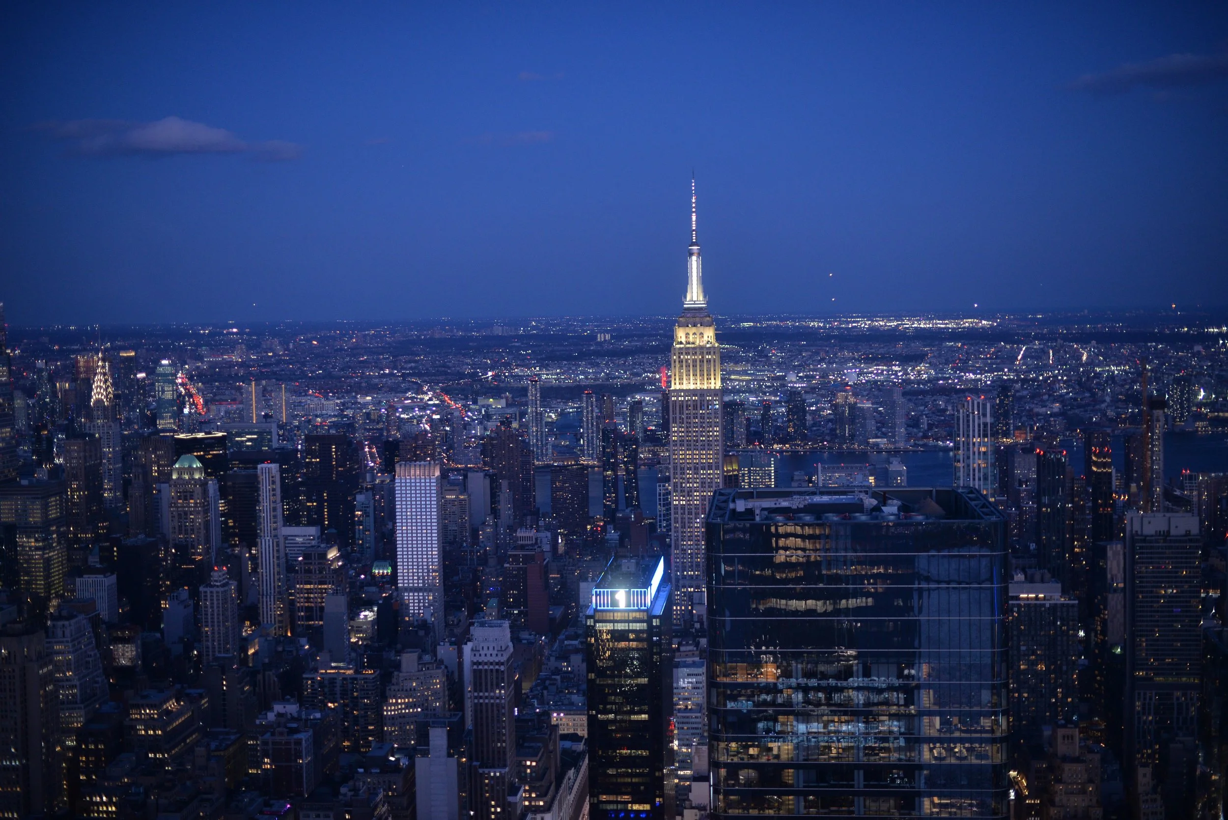 Nighttime aerial view of Manhattan skyline with Empire State Building illuminated and city lights reflecting on the water.