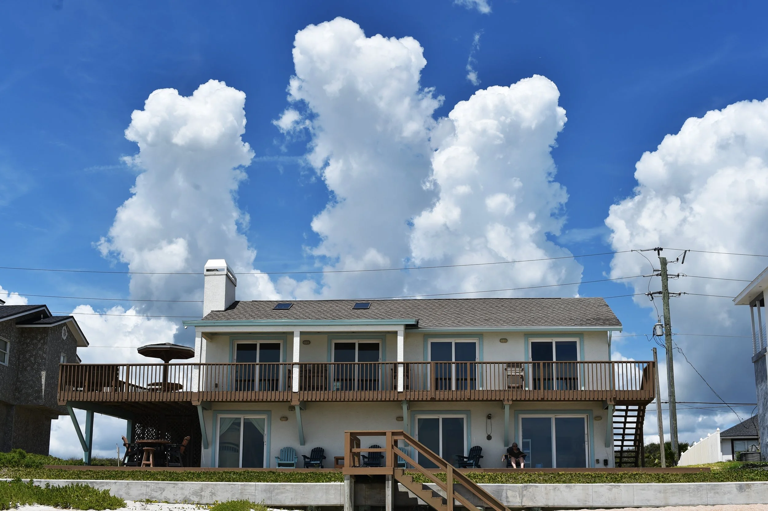 Backyard view of a two-story house with a deck, chairs, and a person sitting, under a blue sky with large white clouds, power lines, and neighboring houses visible.