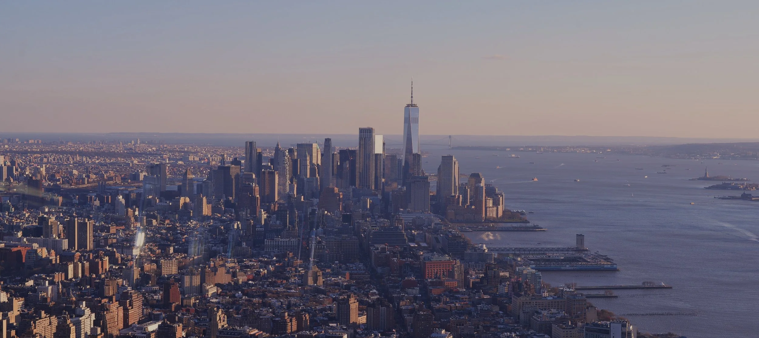 Aerial view of Manhattan skyline in New York City with tall skyscrapers, including One World Trade Center, near the waterfront, with water and boats in the background during daytime.