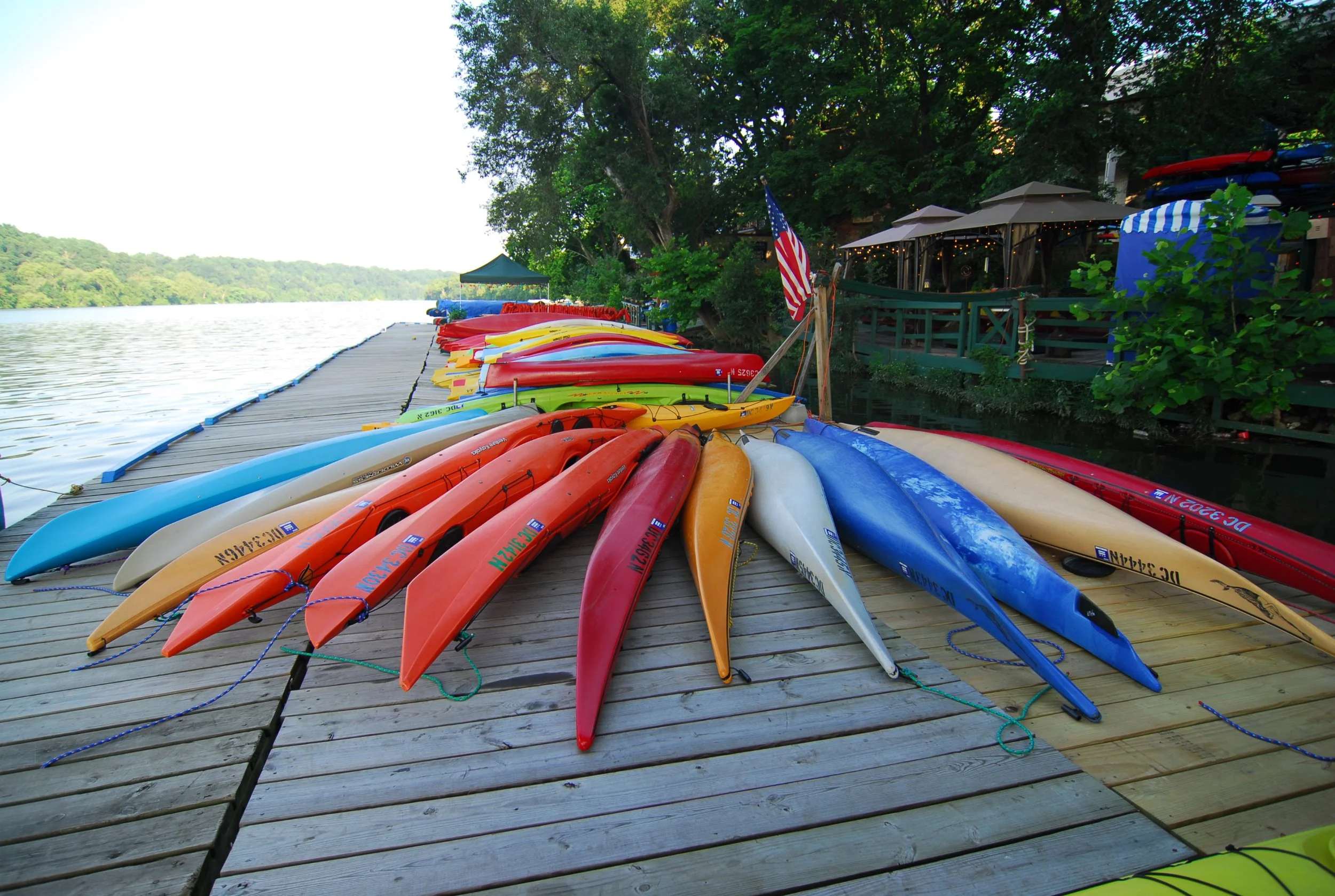 Colorful kayaks lined up on a dock beside a body of water, with a wooded shoreline and a boat house or restaurant in the background.