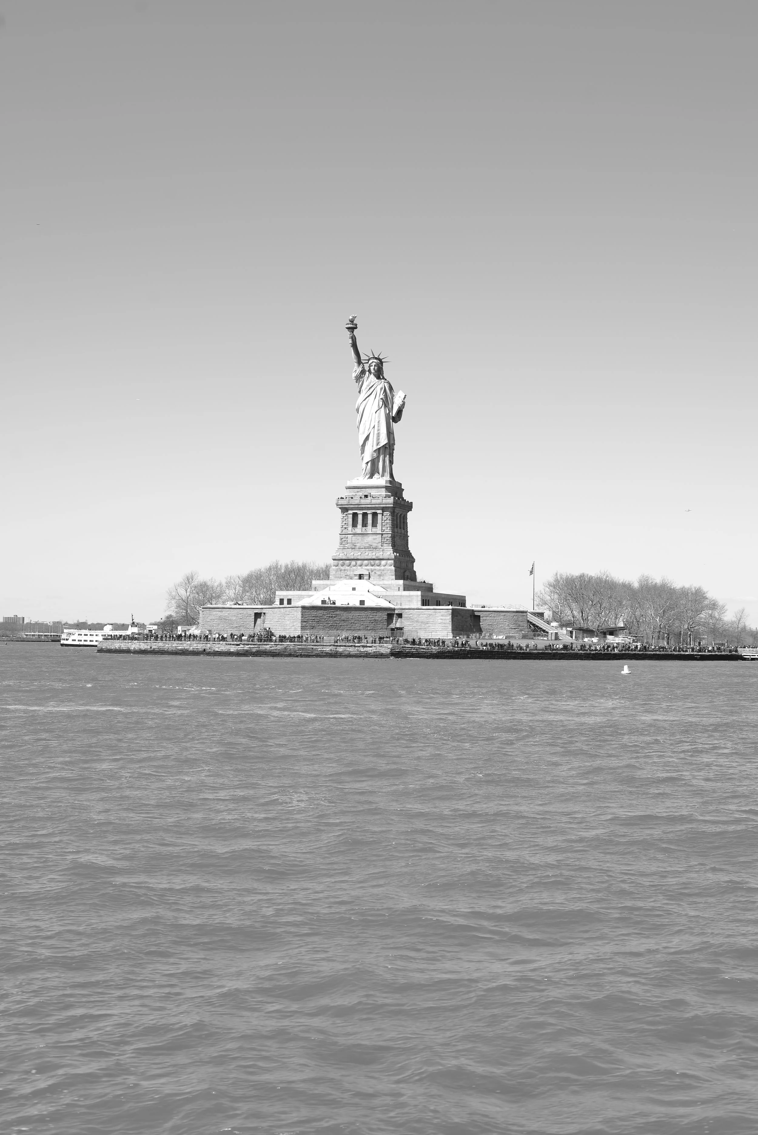 Black and white photo of the Statue of Liberty standing on a pedestal with a body of water in the foreground and trees in the background.