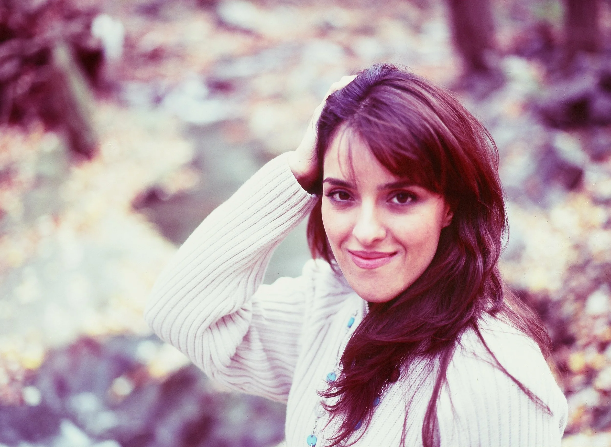 A young woman with long, wavy reddish-brown hair smiling and posing outdoors against a blurred background of trees with fall foliage. She is wearing a light-colored, long-sleeved top and has one arm raised, touching her hair.