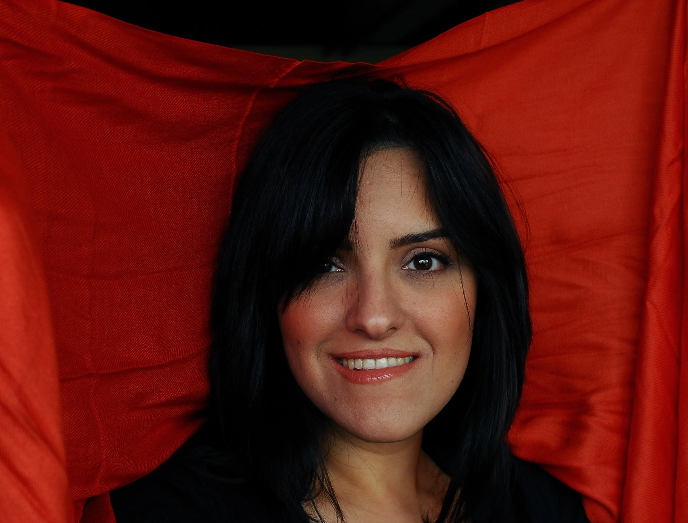 A woman smiling with black hair, partially covered by a red fabric overhead.
