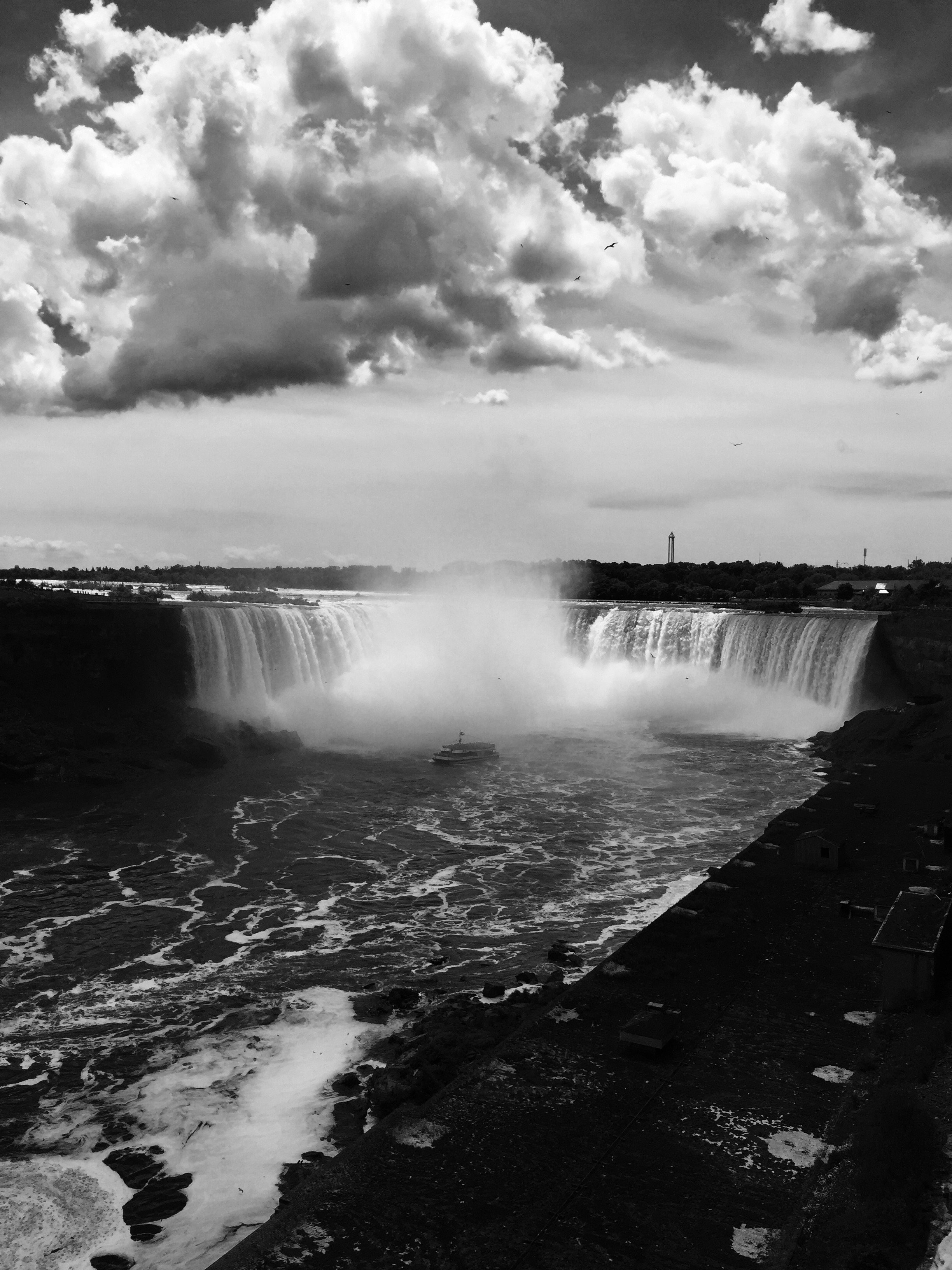 Black and white photograph of Niagara Falls with clouds in the sky, a boat near the waterfall’s base, and a viewing platform on the right side.