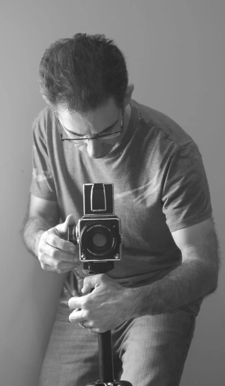 A man with glasses taking a black and white photo with a vintage camera, looking down at the camera.
