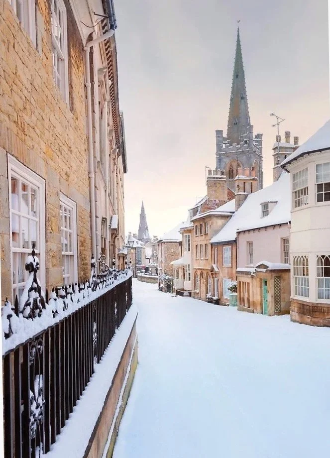 Snow-covered street in a historic neighborhood with stone and brick buildings, a tall church steeple, and a cloudy sky.