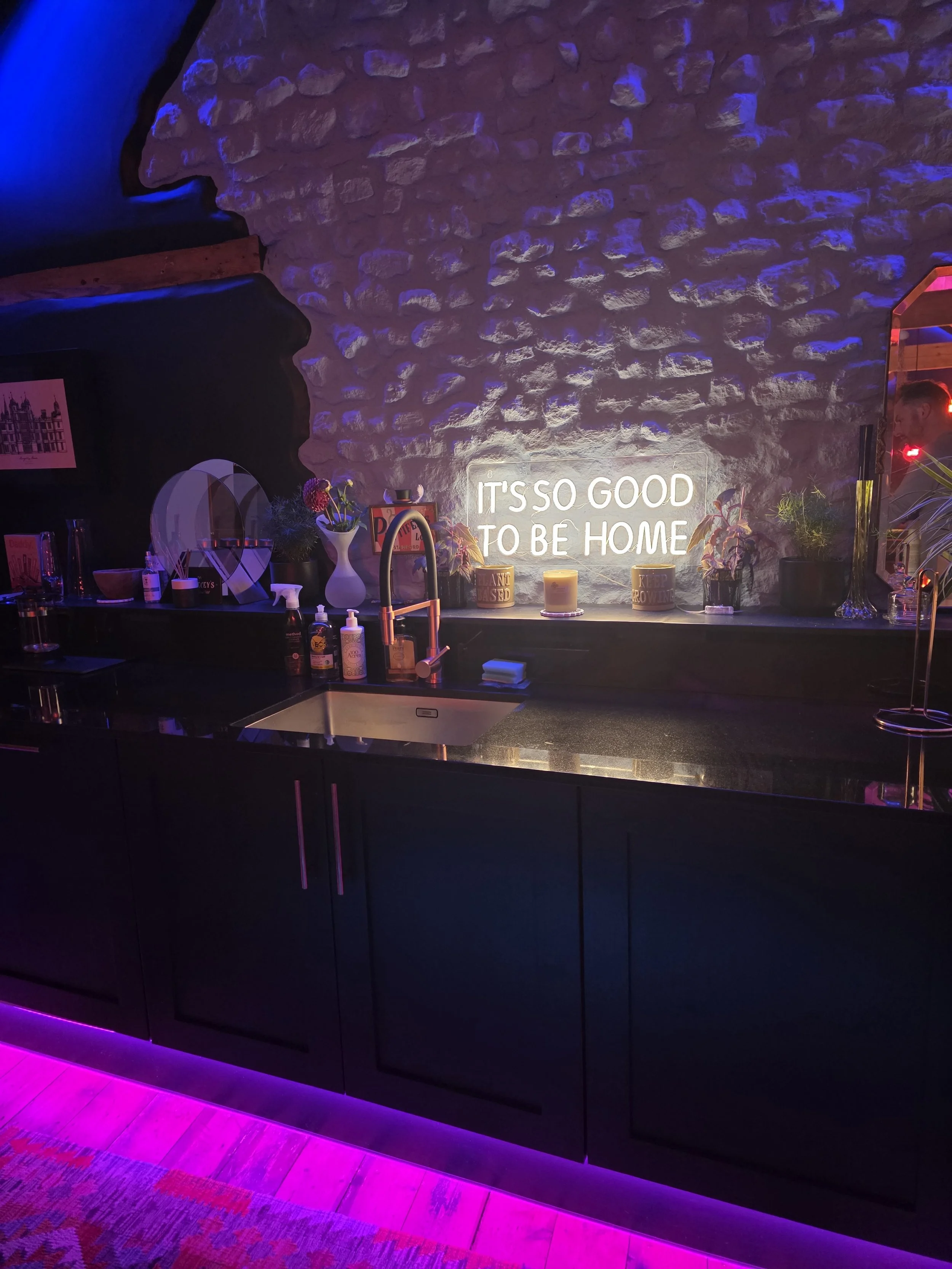 A modern kitchen with dark cabinets, a white sink, and pink and blue accent lighting. Decor includes potted plants, candles, and a neon sign that reads 'IT'S SO GOOD TO BE HOME' on a stone wall.