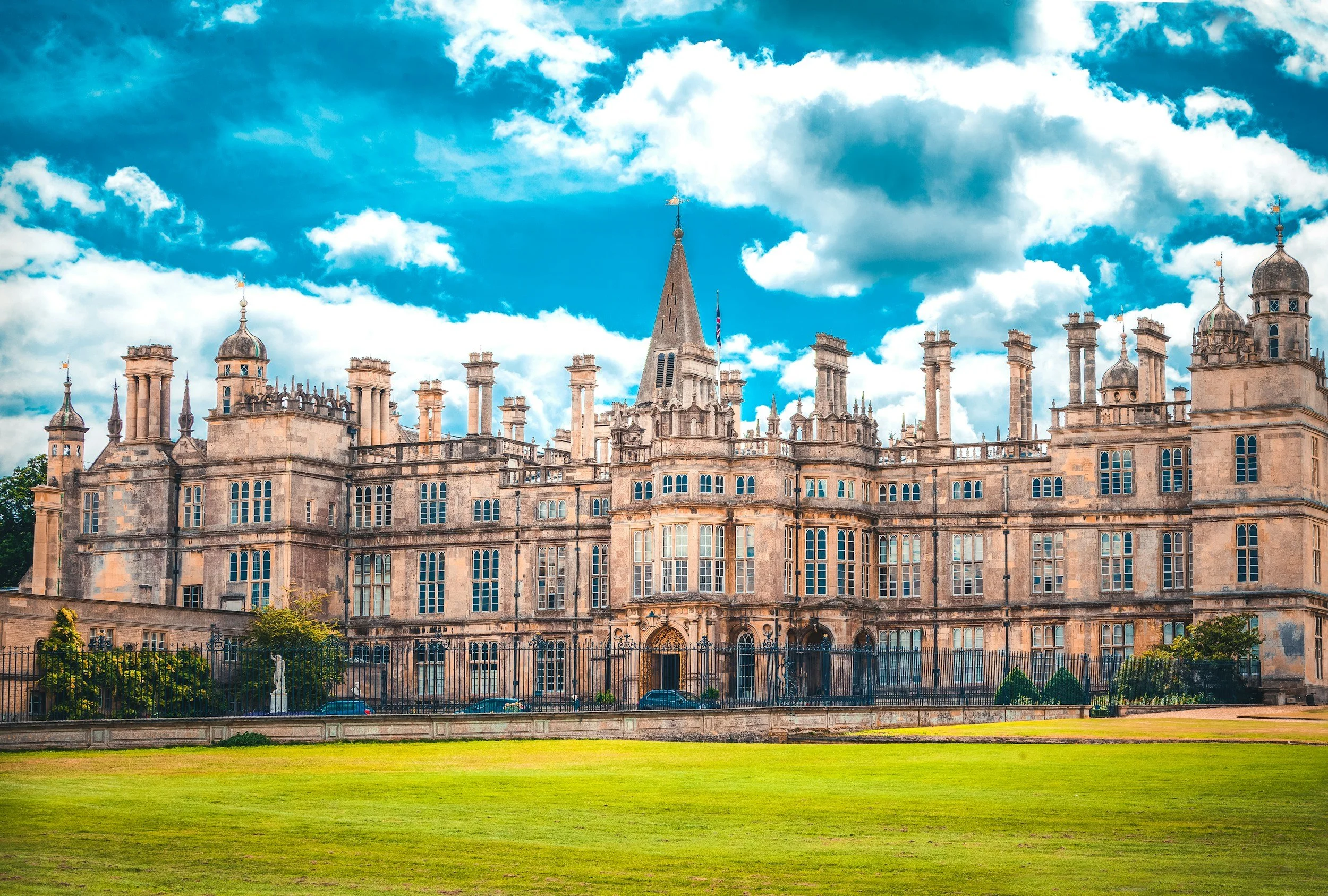 A historic castle with turrets, spires, and chimney stacks behind a black fence, set against a partly cloudy blue sky with a grassy foreground.