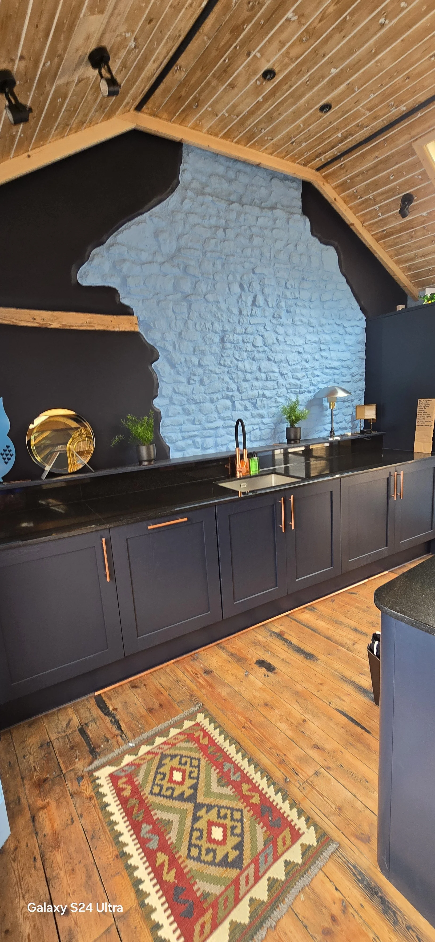 A modern kitchen with dark gray cabinets, black countertop, and a blue brick accent wall. The ceiling is wood-paneled with track lighting. There are small potted plants, a lamp, and decorative items on the counter. A vibrant patterned rug is on a wooden floor.
