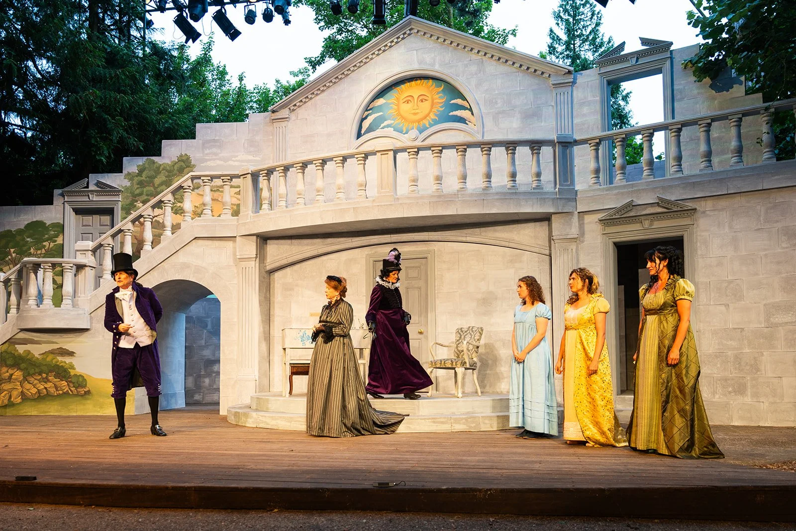 Actors performing on a stage set resembling a classical mansion with a sun and moon mural, featuring six women and one man dressed in vintage clothing.