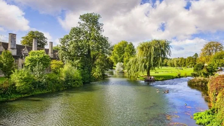 Scenic view of a river flowing past green trees and a historic building with multiple chimneys under a partly cloudy sky.