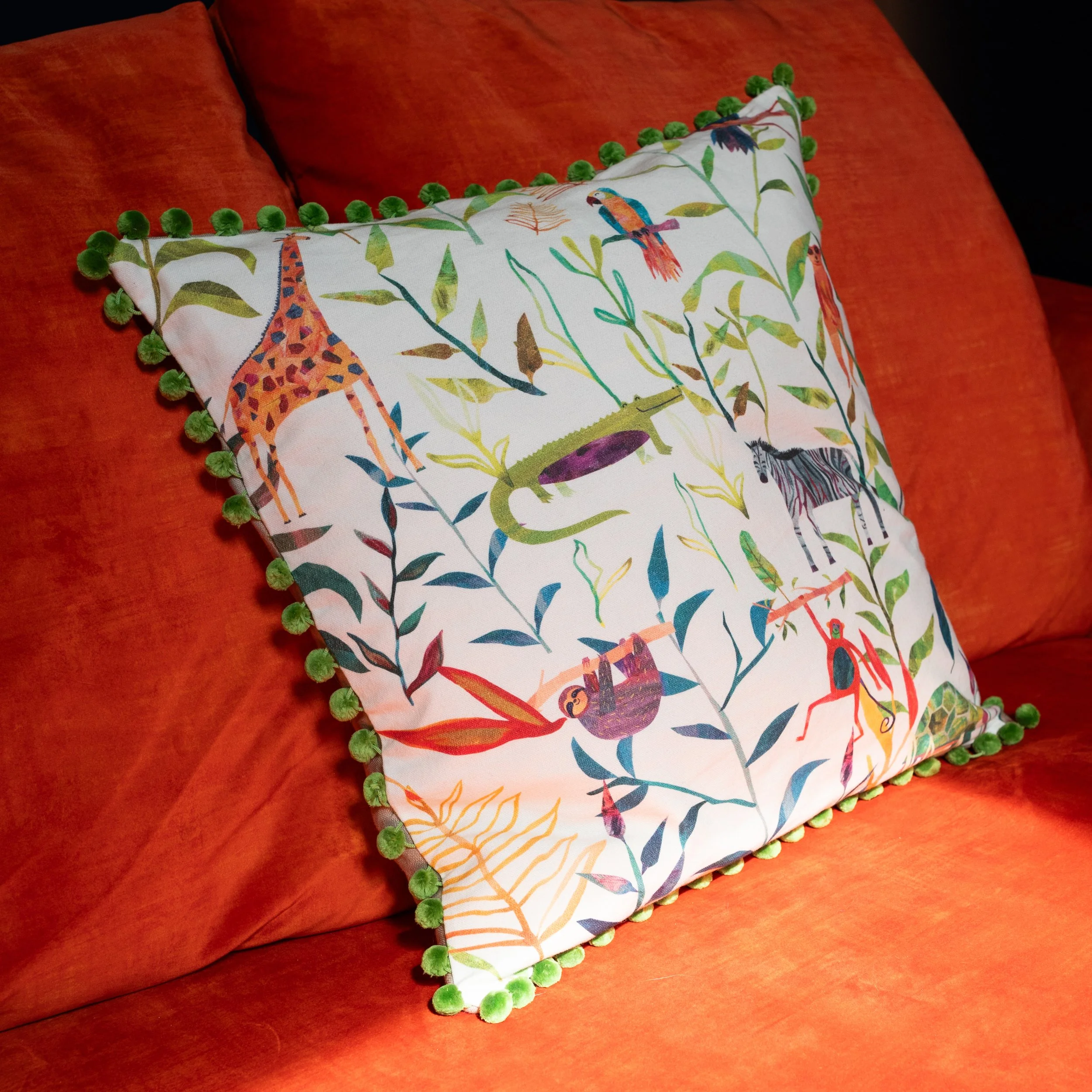 Decorative pillow with multicolored animal and jungle pattern, green pom-pom trim, on a red-orange sofa.