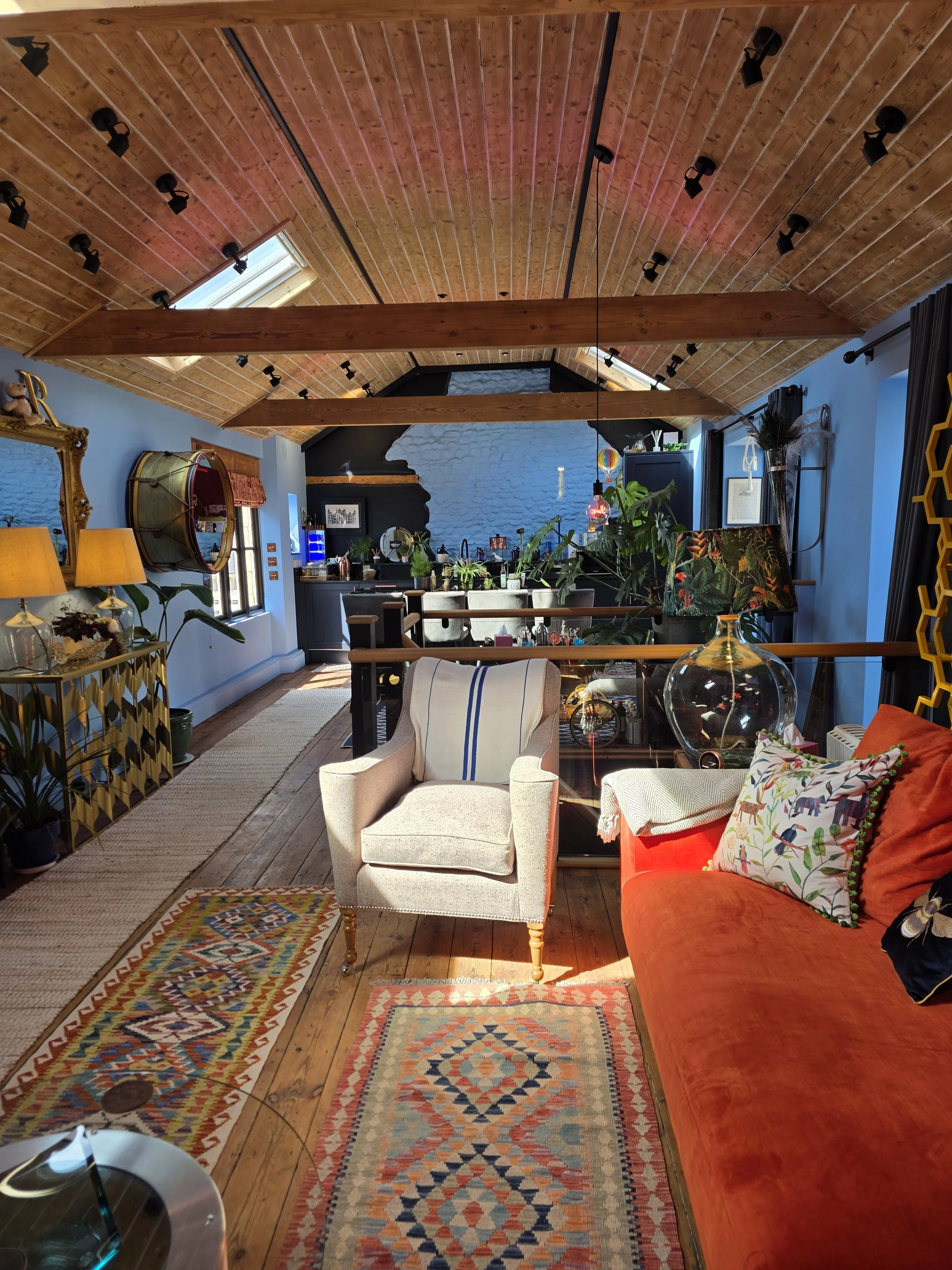 A cozy living room with a wooden ceiling and beams, decorated with colorful rugs and cushions. There are plants, a gold-framed mirror, lamps, and various decorative items, with a partial view of the kitchen in the background.