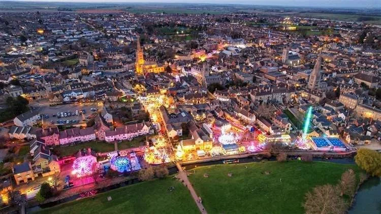 Aerial view of a city at dusk with colorful lights and a fairground with rides, including a Ferris wheel and a tall tower. The cityscape has historic buildings, a river, and green park areas.