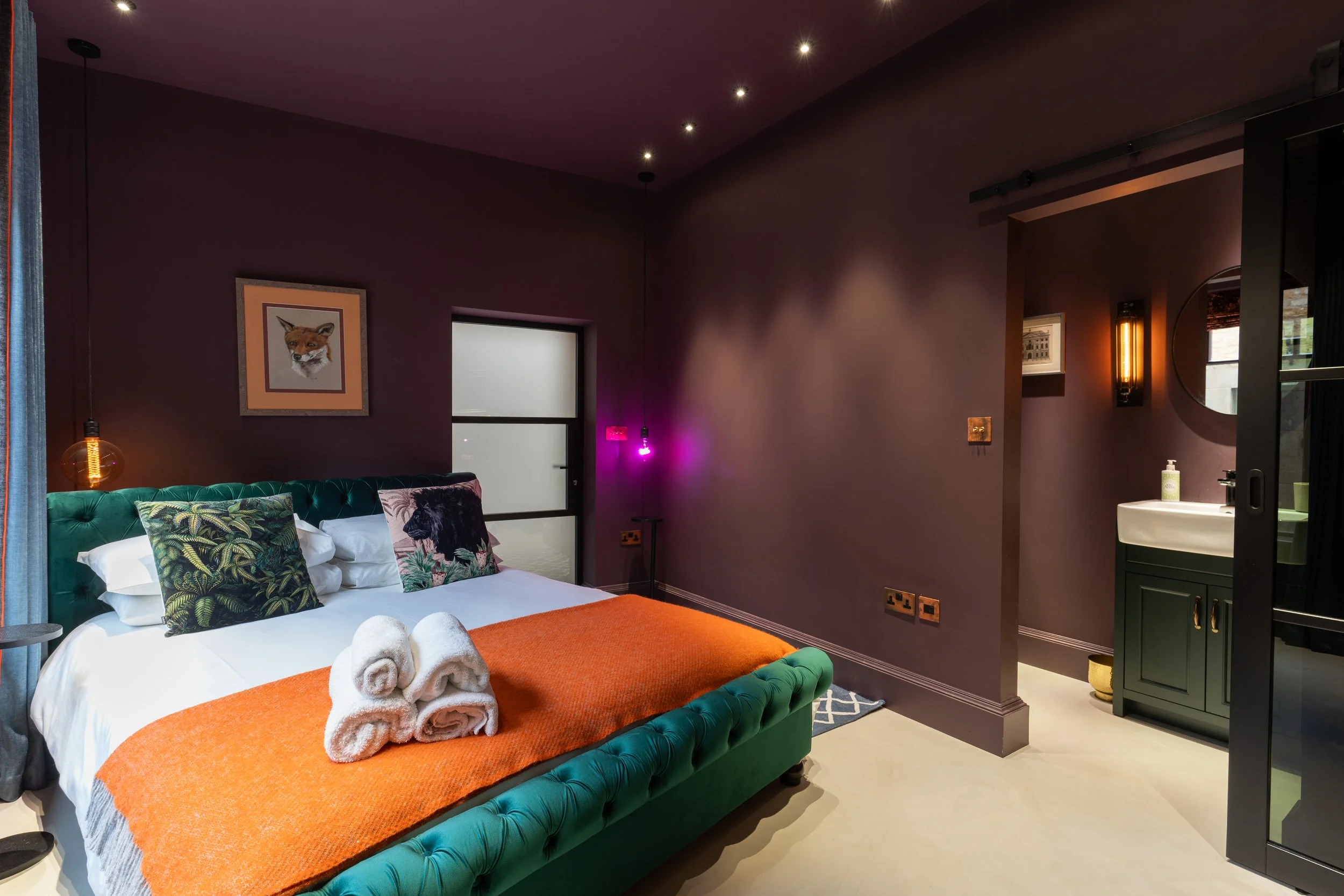 A bedroom with purple walls, a green velvet bed with white and patterned pillows, and an orange throw blanket. Towels are rolled on the bed. There are pendant and wall lights, a window, and artwork, including a framed fox portrait. A small bathroom area with a sink and mirror is visible.