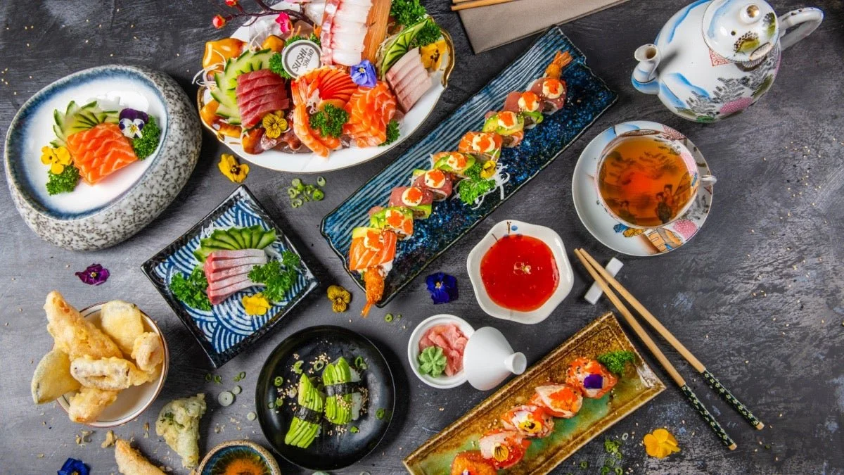 Assorted Japanese sushi and sashimi dishes with chopsticks, soy sauce, tea, and decorative edible flowers on a dark table.