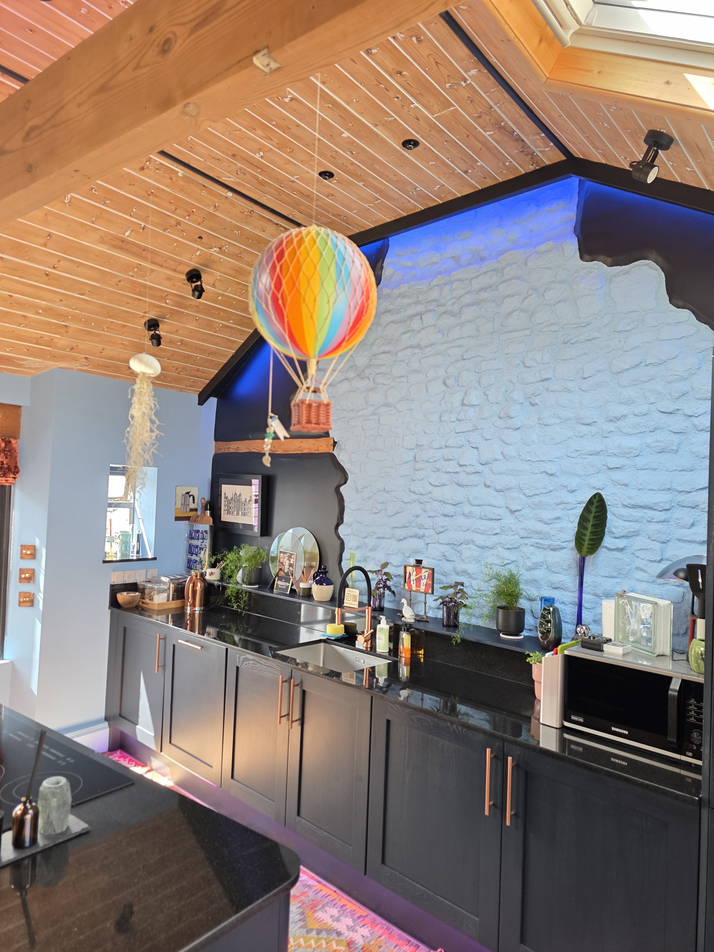 Interior view of a kitchen with a black countertop, gray cabinets with copper handles, a blue distressed wall, and a wooden ceiling with track lighting. There are various kitchen items and plants on the counter.