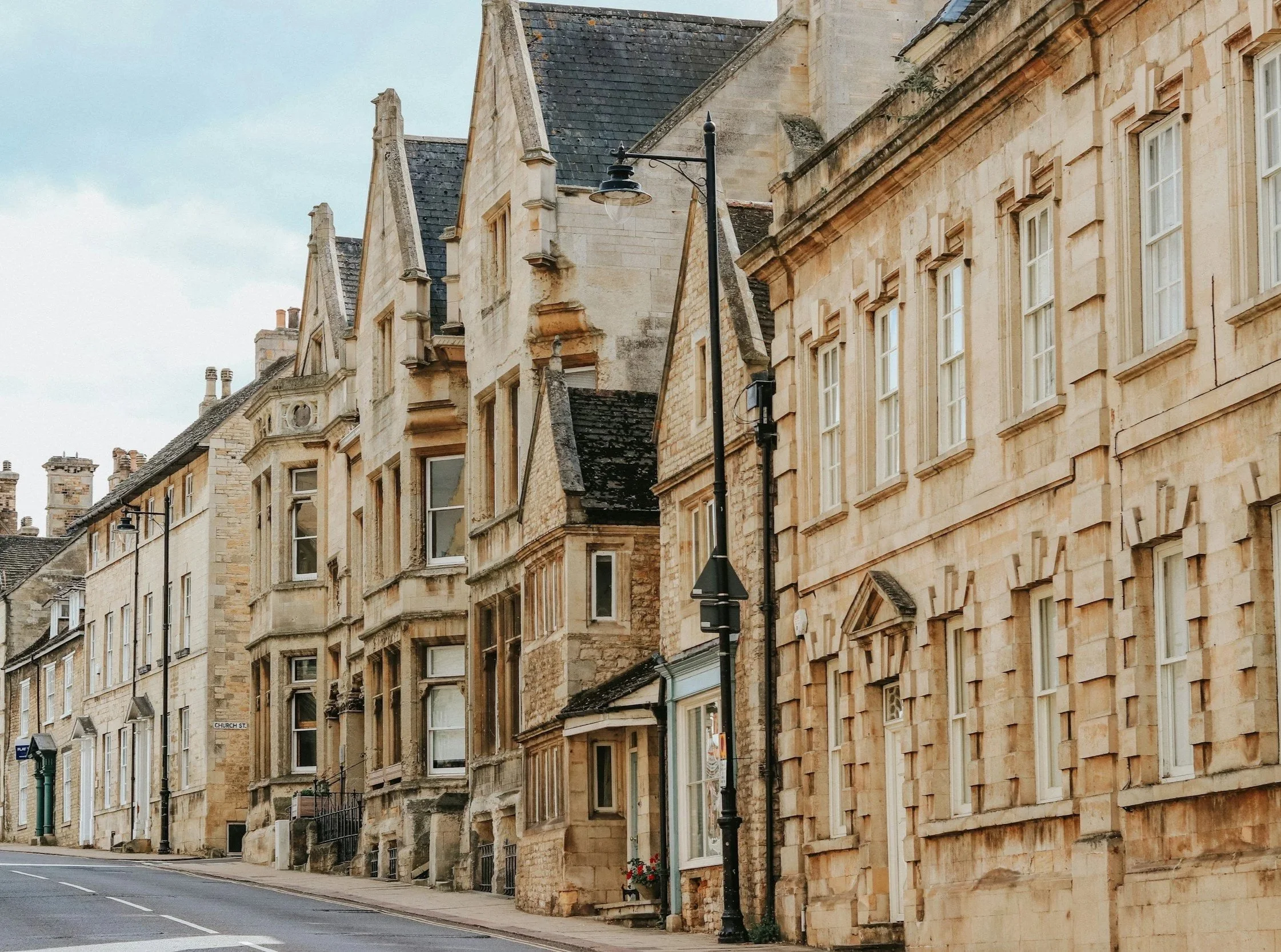 A row of historic stone buildings with large windows and steep rooftops along a quiet street, with street lamps and a cloudy sky.