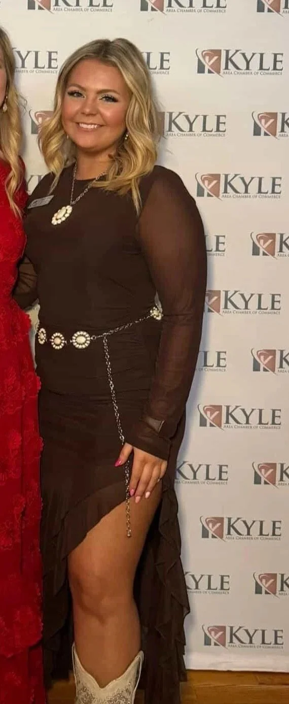 A young woman with blonde hair in loose waves, wearing a long, semi-sheer brown dress with a high slit and western-style boots, standing in front of a backdrop with the Kyle Chamber of Commerce logo.