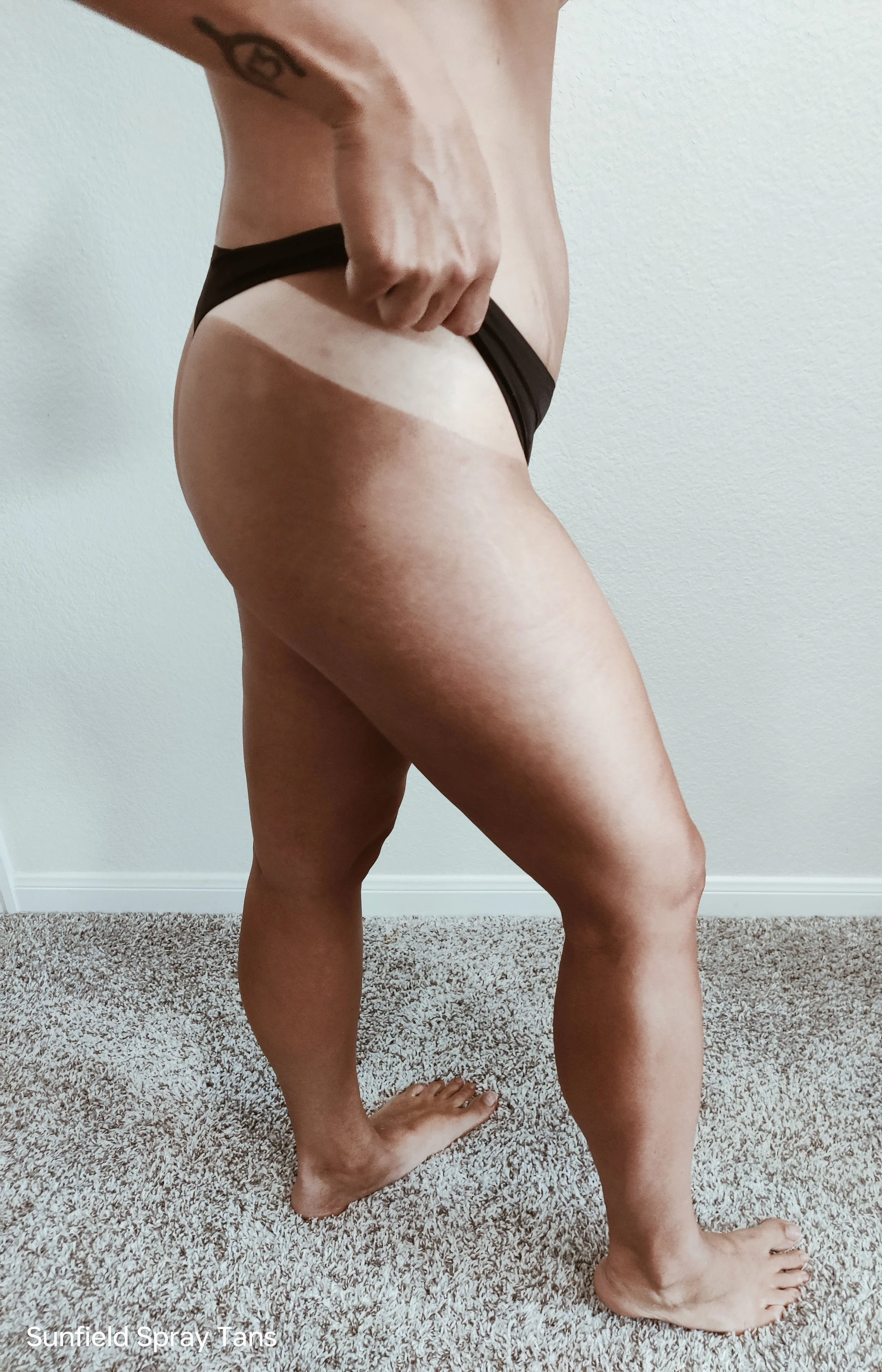 Side view of a person standing indoors, wearing black underwear, with visible tan lines from sun exposure.