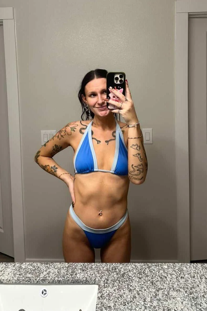 A woman taking a mirror selfie in a bathroom, wearing a blue and white bikini, with tattoos on her arms, and a belly button piercing.