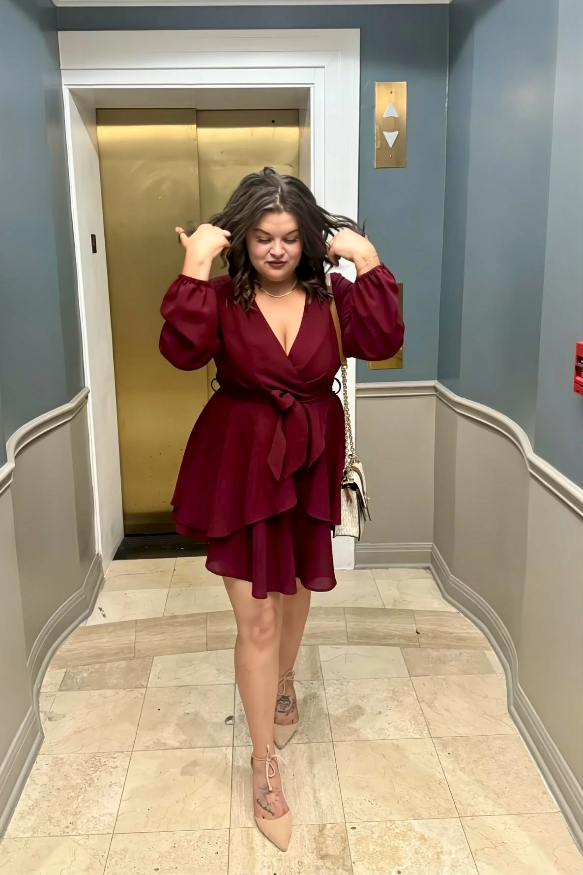 A woman with dark hair in loose waves, wearing a deep red dress with puffed sleeves and a waist tie, stands in front of an elevator, adjusting her hair and smiling softly.
