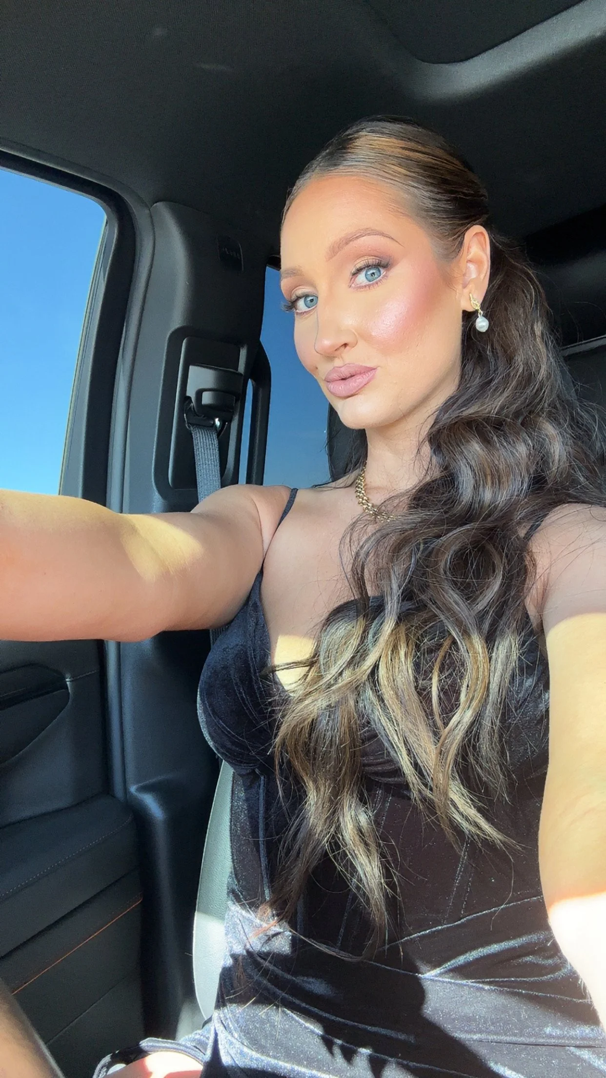 A woman taking a selfie inside a vehicle, with a clear blue sky visible through the window. She has long, wavy brown hair, blue eyes, and is wearing makeup, jewelry including earrings and a necklace, and a black sleeveless top.