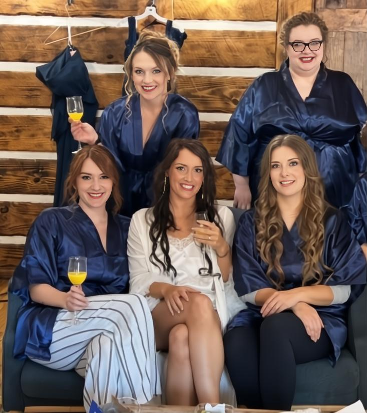 Five women in navy-blue satin robes, sitting and standing in a rustic wooden room, with two women holding glasses of mimosas, celebrating or preparing for a wedding.
