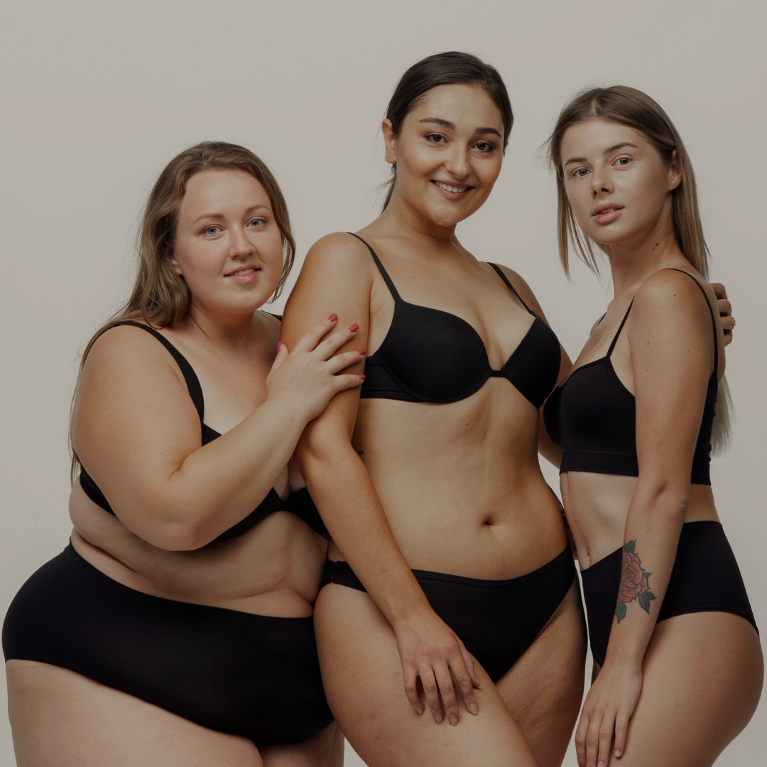 Three women standing together, wearing black bras and underwear, smiling and posing against a plain, light-colored background.