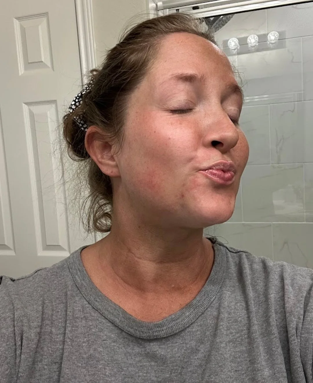 A woman with her eyes closed, pursing her lips, with her hair tied back and wearing a gray t-shirt, in a bathroom with a mirror and tiled wall in the background.