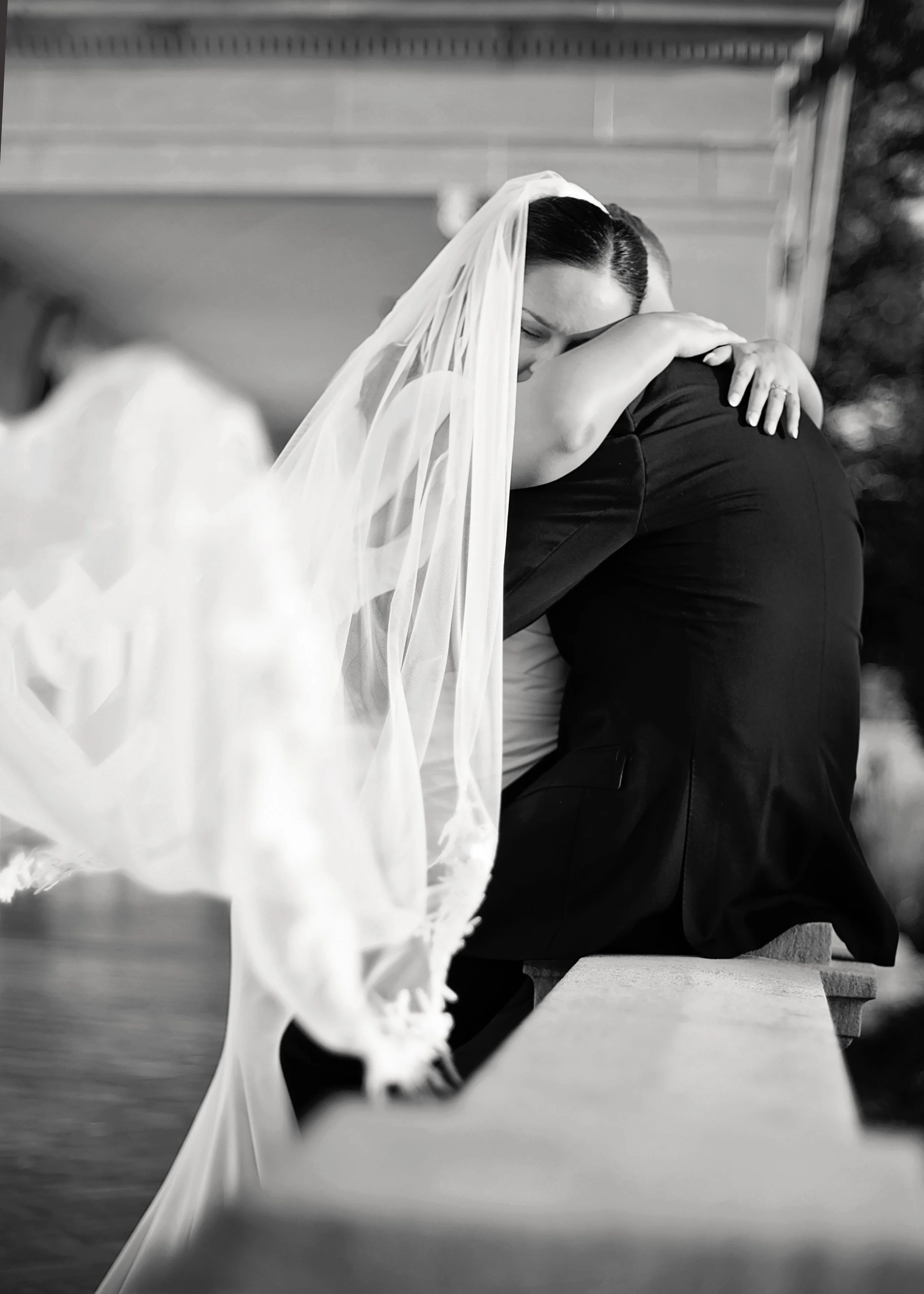 Bride in a stunning off the shoulder gown posing with groom against an elegant stone mansion.