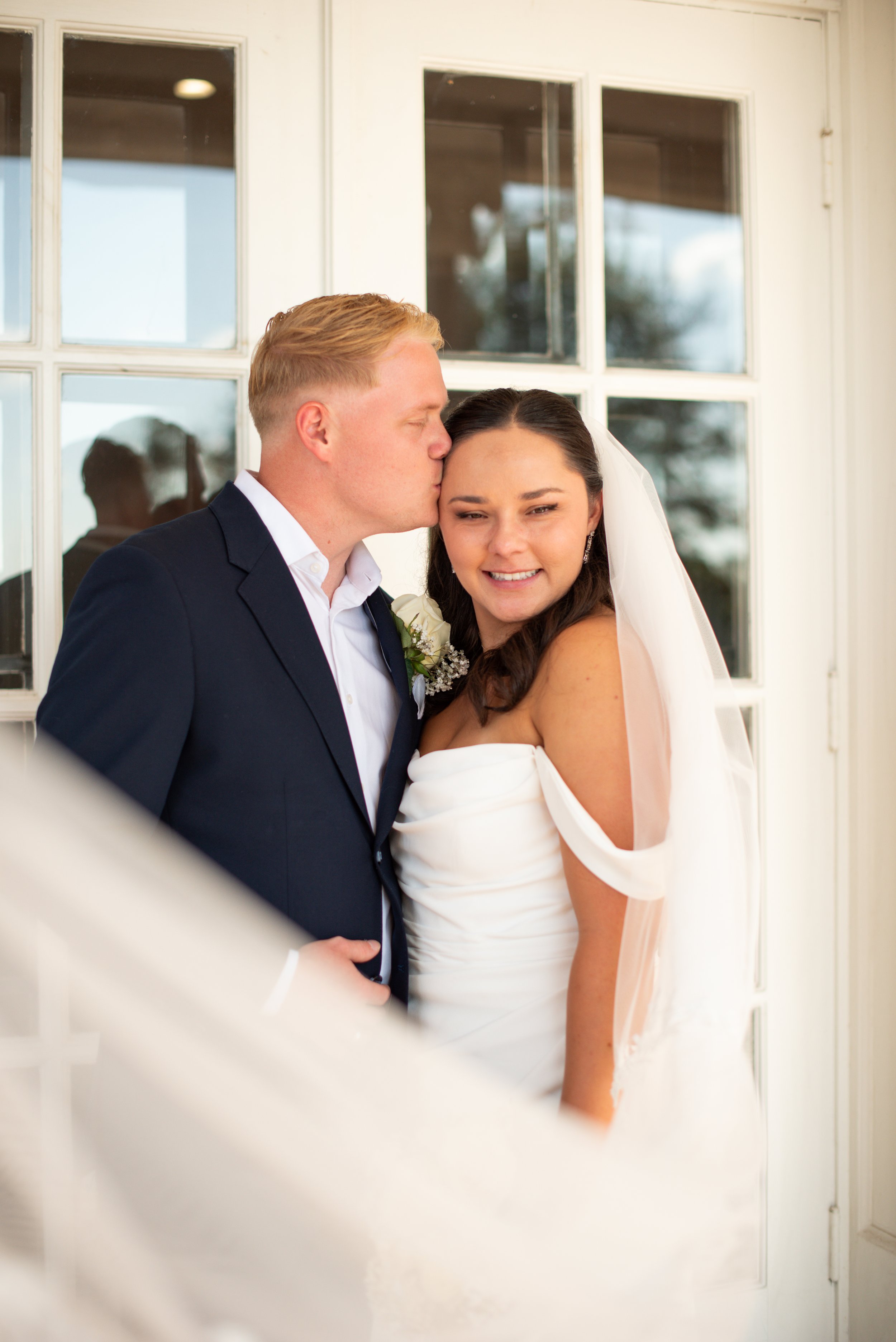 Bride in a stunning off the shoulder gown posing with groom against an elegant stone mansion.