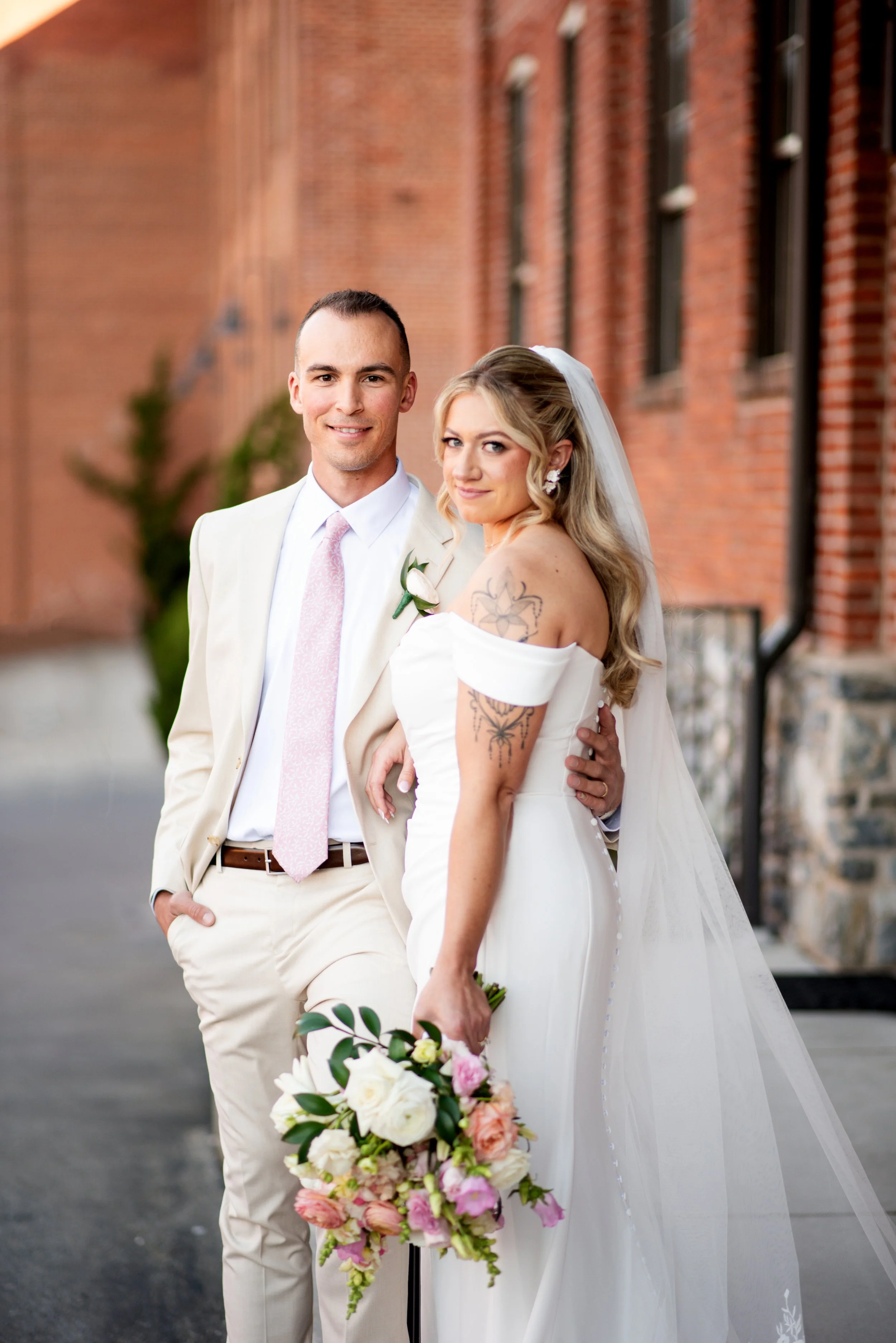 A bride and groom standing outdoors in front of a brick building, celebrating their wedding day. The groom is wearing a light-colored suit with a pink tie and the bride is holding a bouquet of pink and white flowers, dressed in a white wedding gown with a veil.